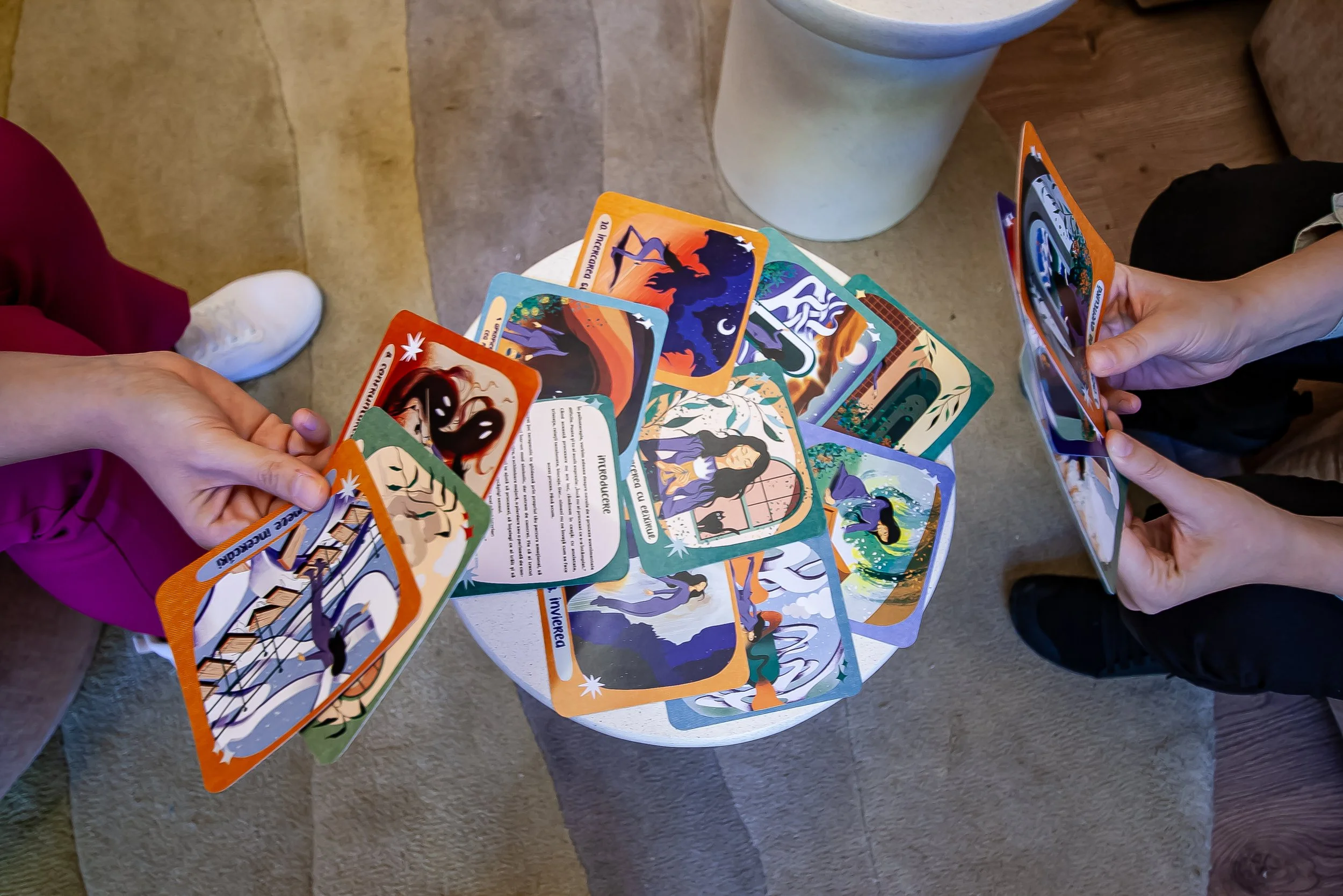 Two people holding and looking at illustrated cards over a small table filled with additional cards, with a white round table and a beige floor in the background. - Carduri Calatoria Eroinei