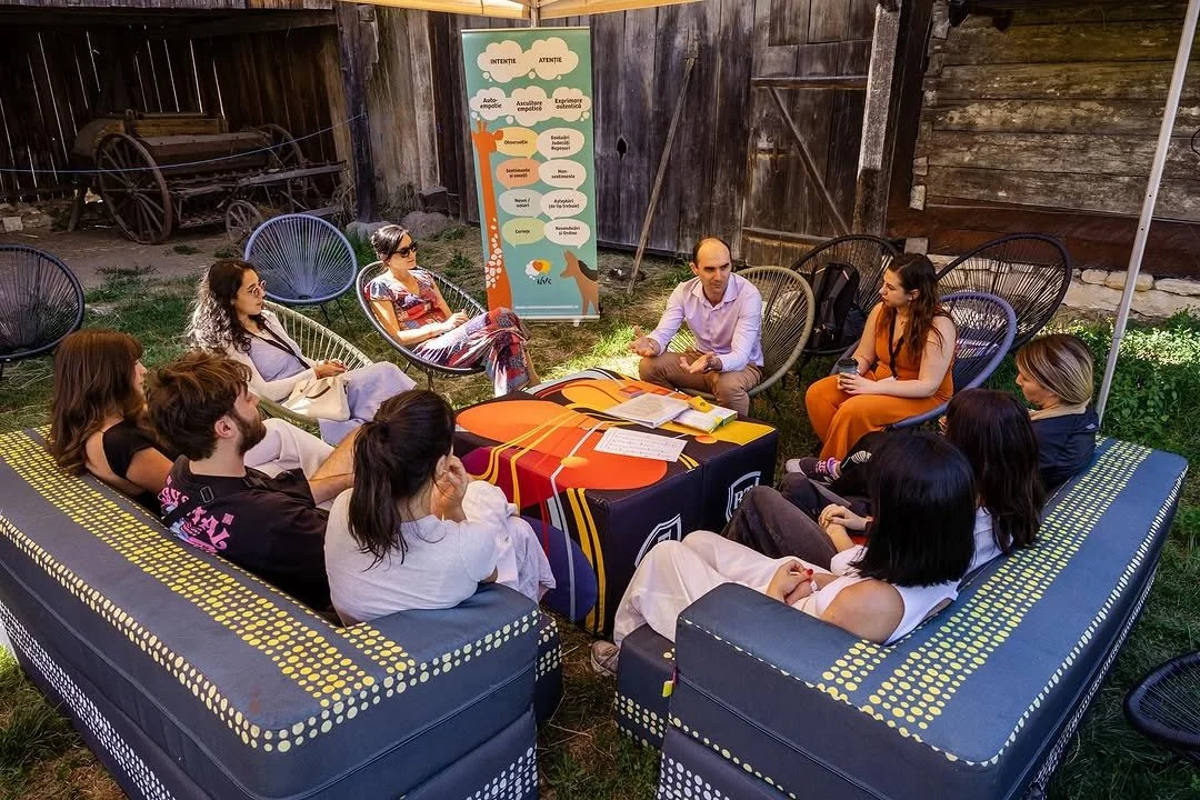 A group of ten people seated in a semi-circle outdoors on various chairs and couches, attending a discussion or meeting, with a table in the center holding books and papers, and a colorful poster with text behind them.