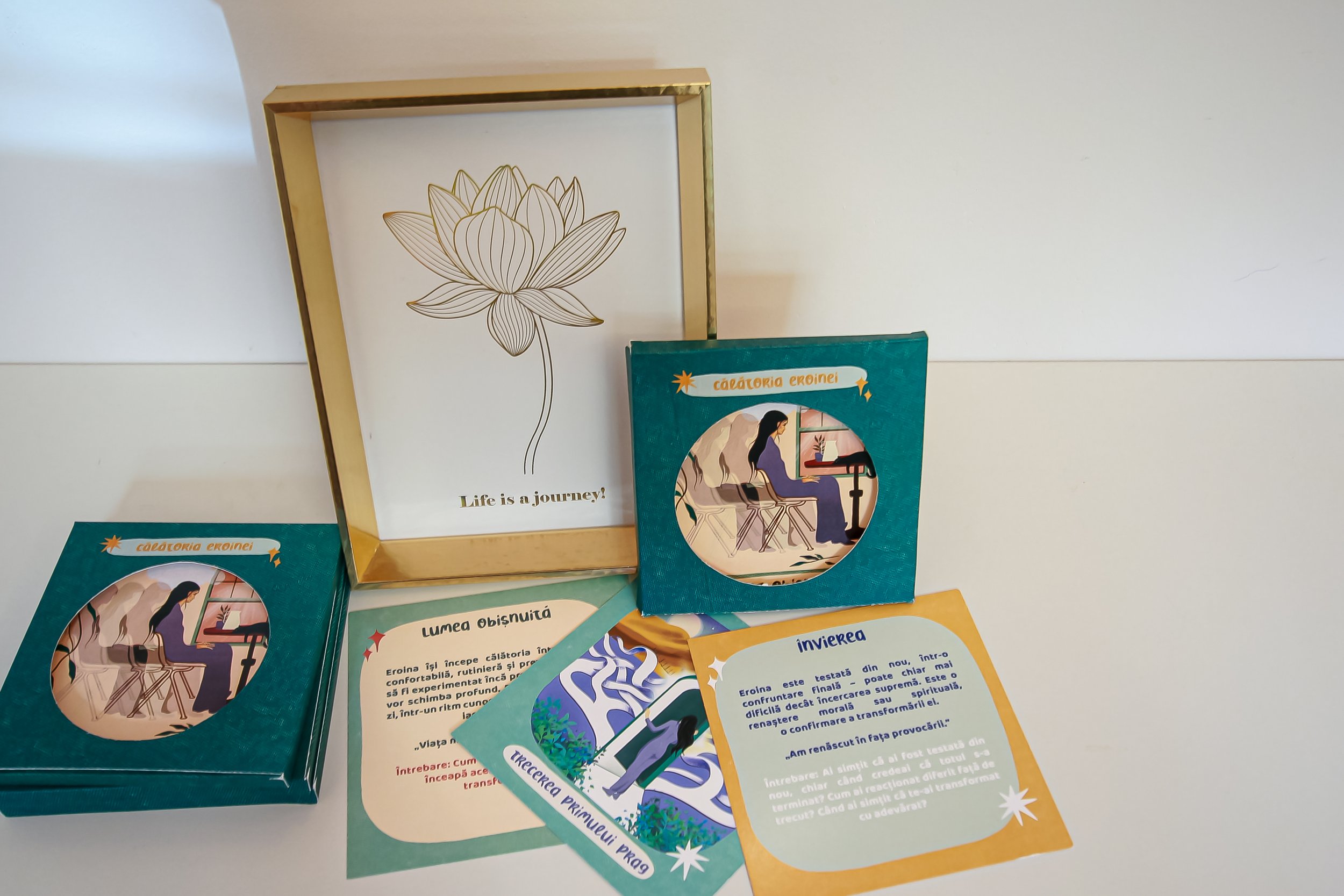 Decorative framed print with a gold border and a line drawing of a lotus flower, and the quote 'Life is a journey?'. Several colorful cards and small boxes with illustrations and text are arranged in front of it. - Carduri Calatoria Eroinei