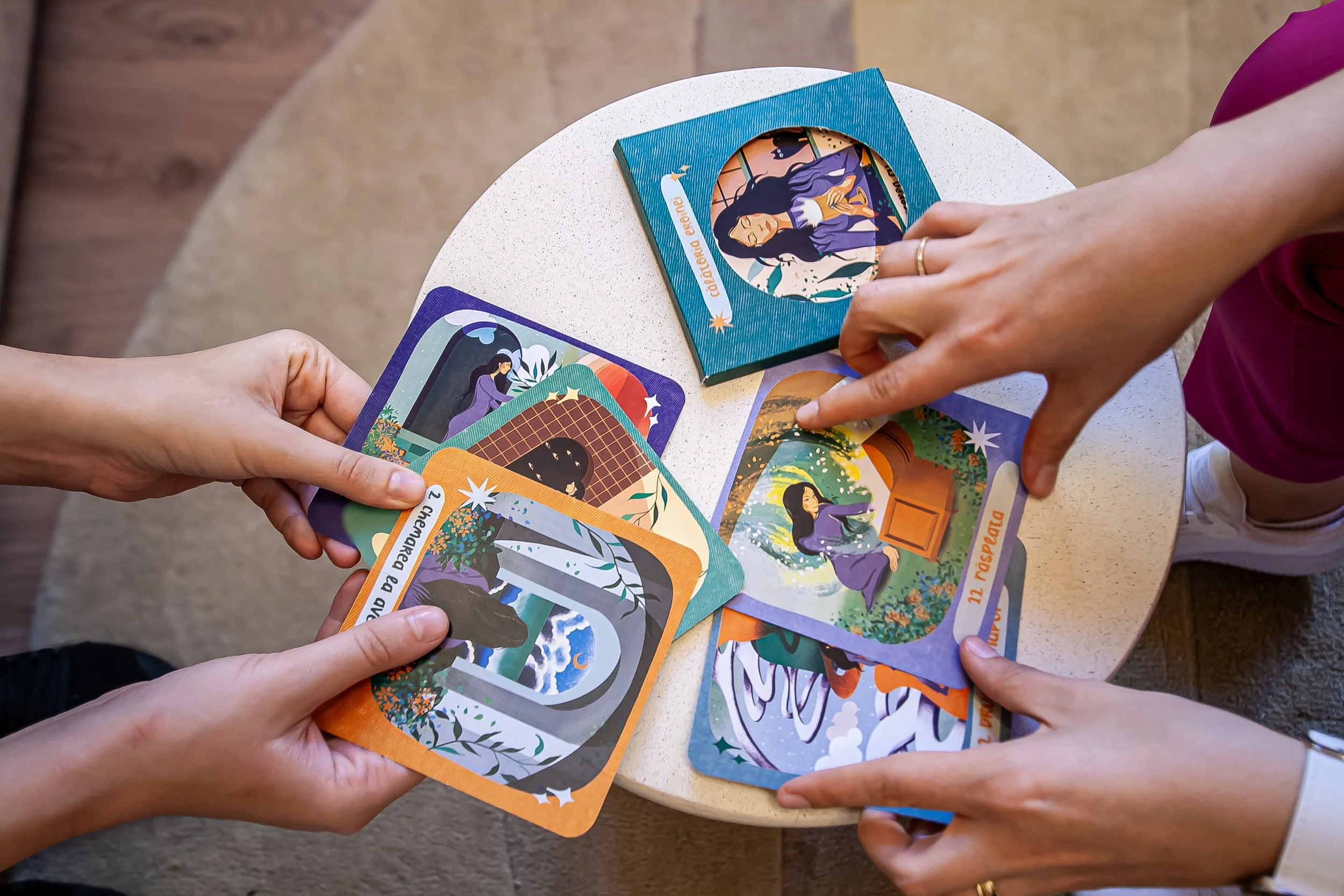 Multiple illustrated cards being shown and discussed by two people on a small round table. - Carduri Calatoria Eroinei