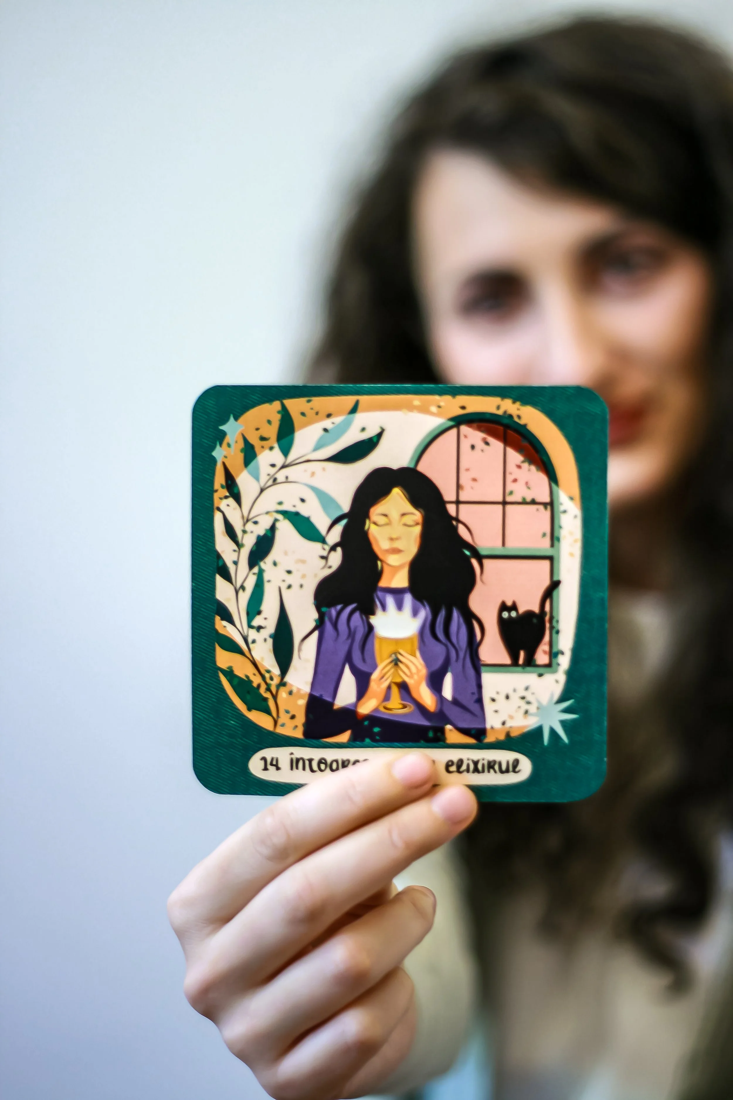 A person holding a colorful illustrated card featuring a woman with long dark hair, holding a glass of drink, with a window and a black cat in the background. - Carduri Calatoria Eroinei