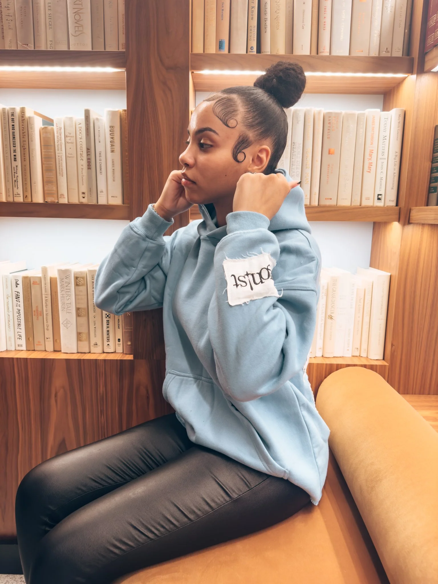 A young woman with a curly ponytail sitting on an orange cushion in front of a wooden bookshelf filled with books, wearing a light blue hoodie and black leather pants, adjusting her hoodie straps.
