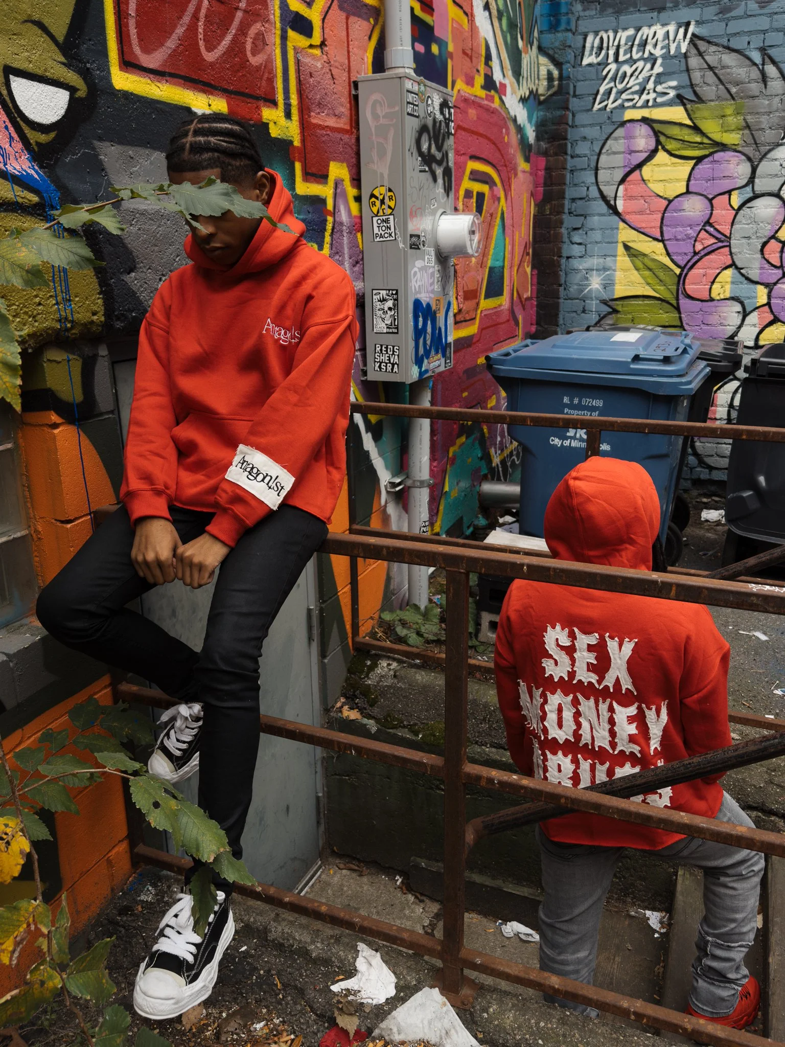 Two people in red hoodies sitting on a metal railing in front of a graffiti-covered wall. One person is sitting with legs crossed, looking down, with leaves covering part of their face. The other has their back to the camera, and the hoodie says 'SEX MONEY BILLS' on it.