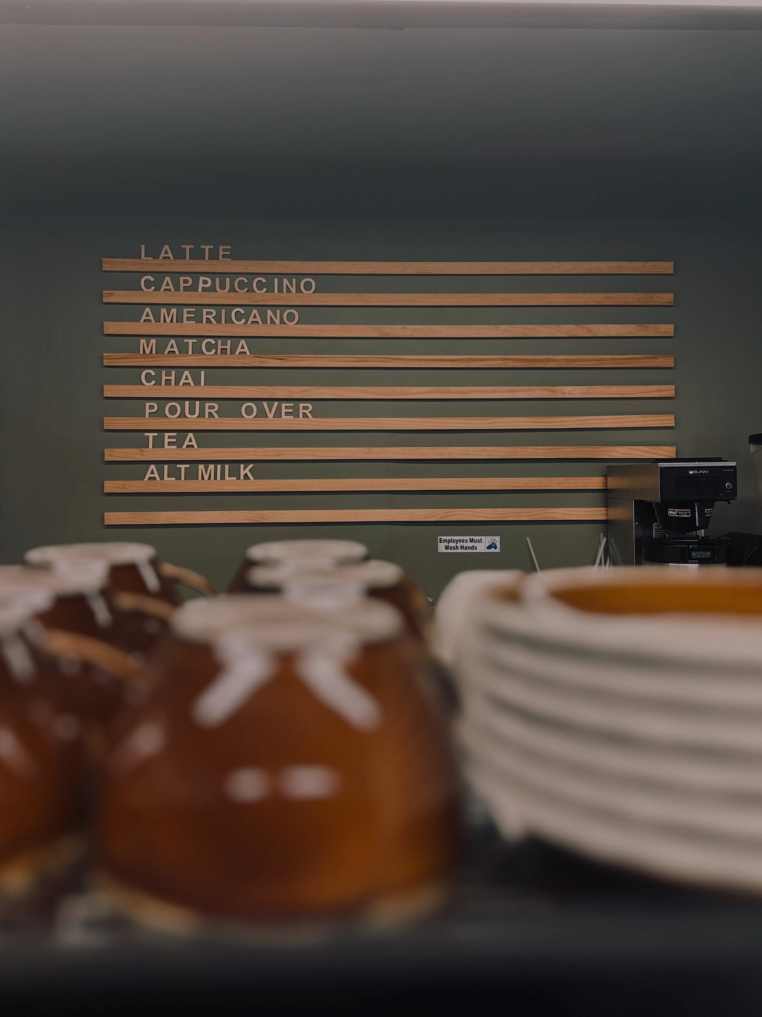 A menu with wooden slats listing coffee drinks and items, with blurred brown mugs and white plates in the foreground; a coffee machine on the right with a small sign below reading "Employees Must Wash Hands."