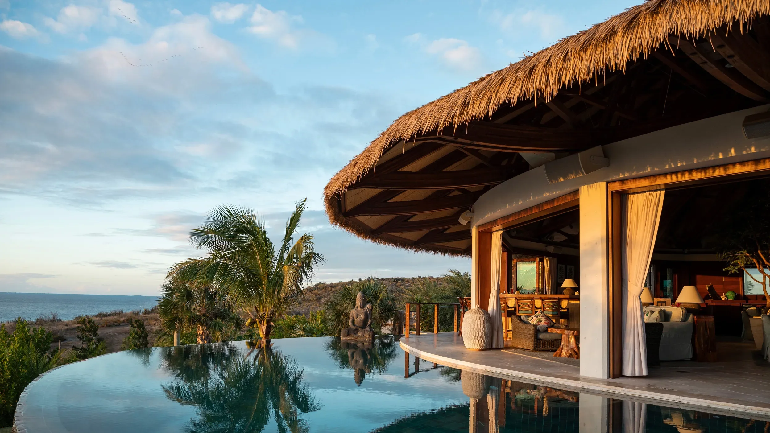 A luxurious beachfront villa with thatched roof, large open windows, and an infinity pool reflecting the sky, overlooking the ocean and surrounding tropical trees during sunset.