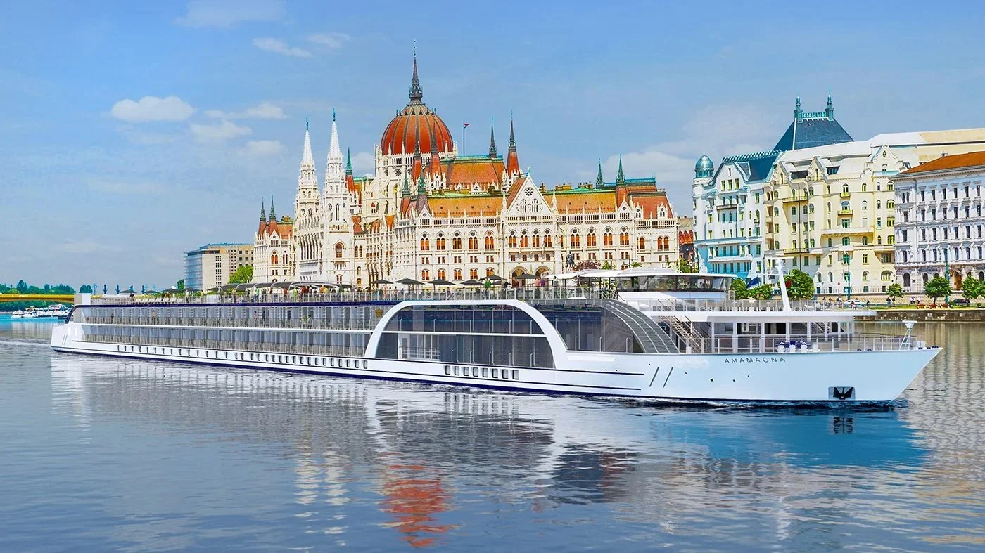 A modern cruise ship named AMANAGNA floating on a river with historic European buildings in the background, under a partly cloudy sky.