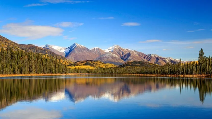 Scenic landscape of snow-capped mountains reflecting in a lake with a forested shoreline under a blue sky.