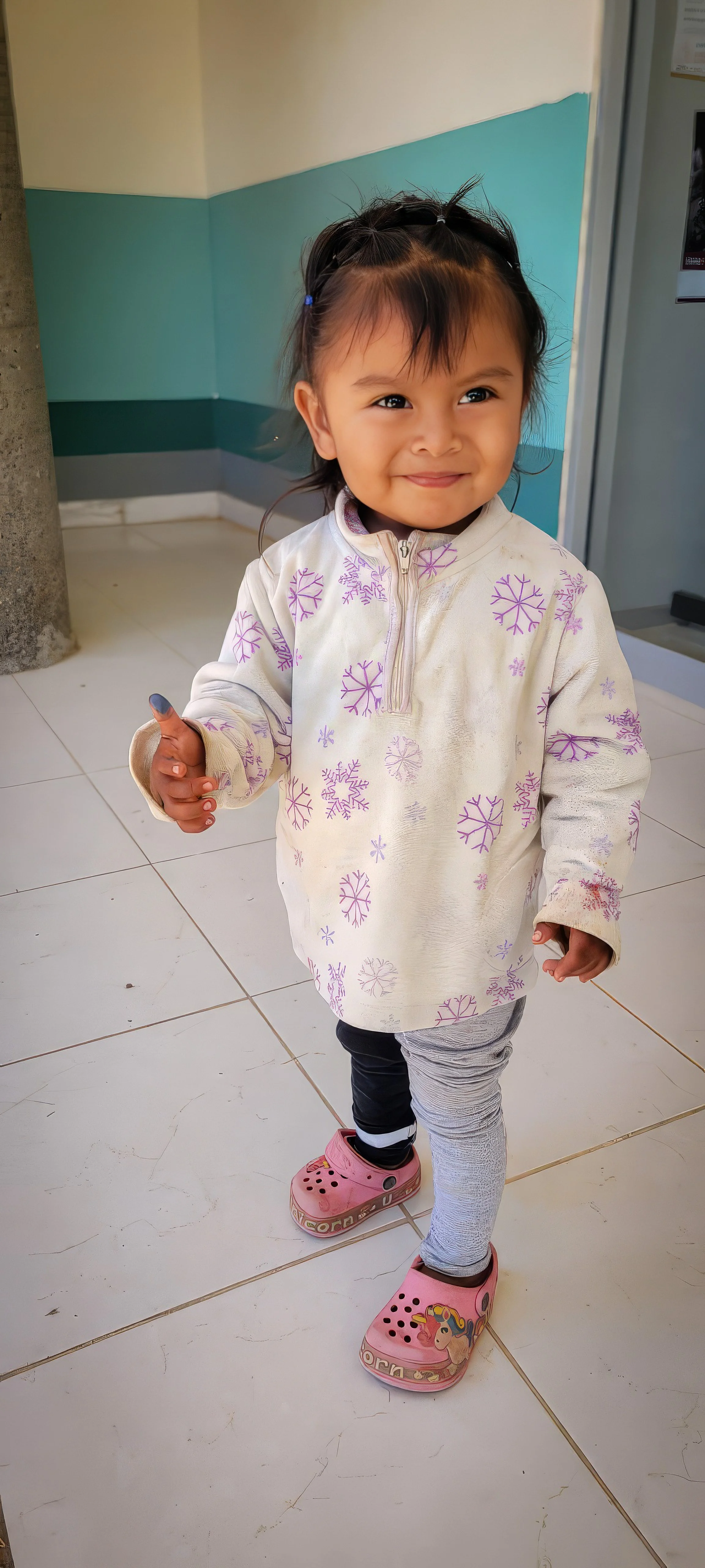 A young girl standing indoors on a tiled floor, smiling and giving a thumbs-up. She is wearing a white jacket with purple snowflake patterns, gray leggings, and pink shoes with cartoon characters.