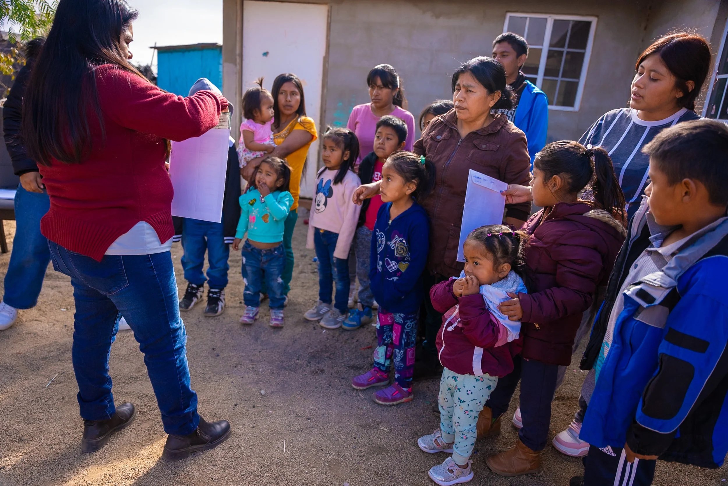 A group of children and adults gathered outdoors, listening to a woman with long hair and wearing a red sweater, who is giving a family their birth certificates. The setting includes weathered buildings and a sunny sky.