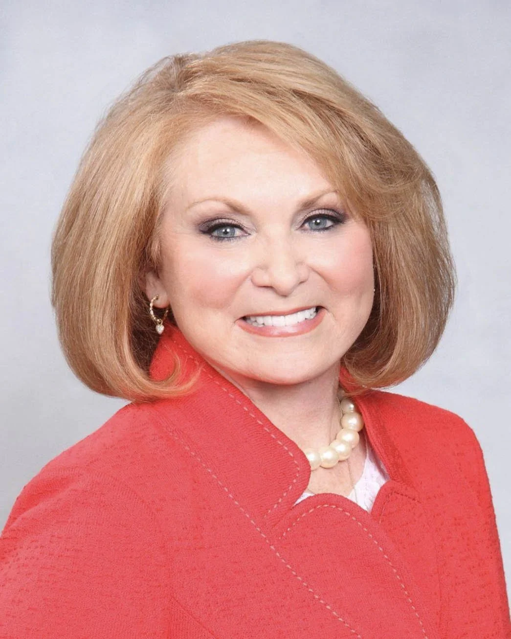 A woman with curly black hair, glasses, and hoop earrings smiling at the camera, wearing a white striped blouse with layered necklaces.