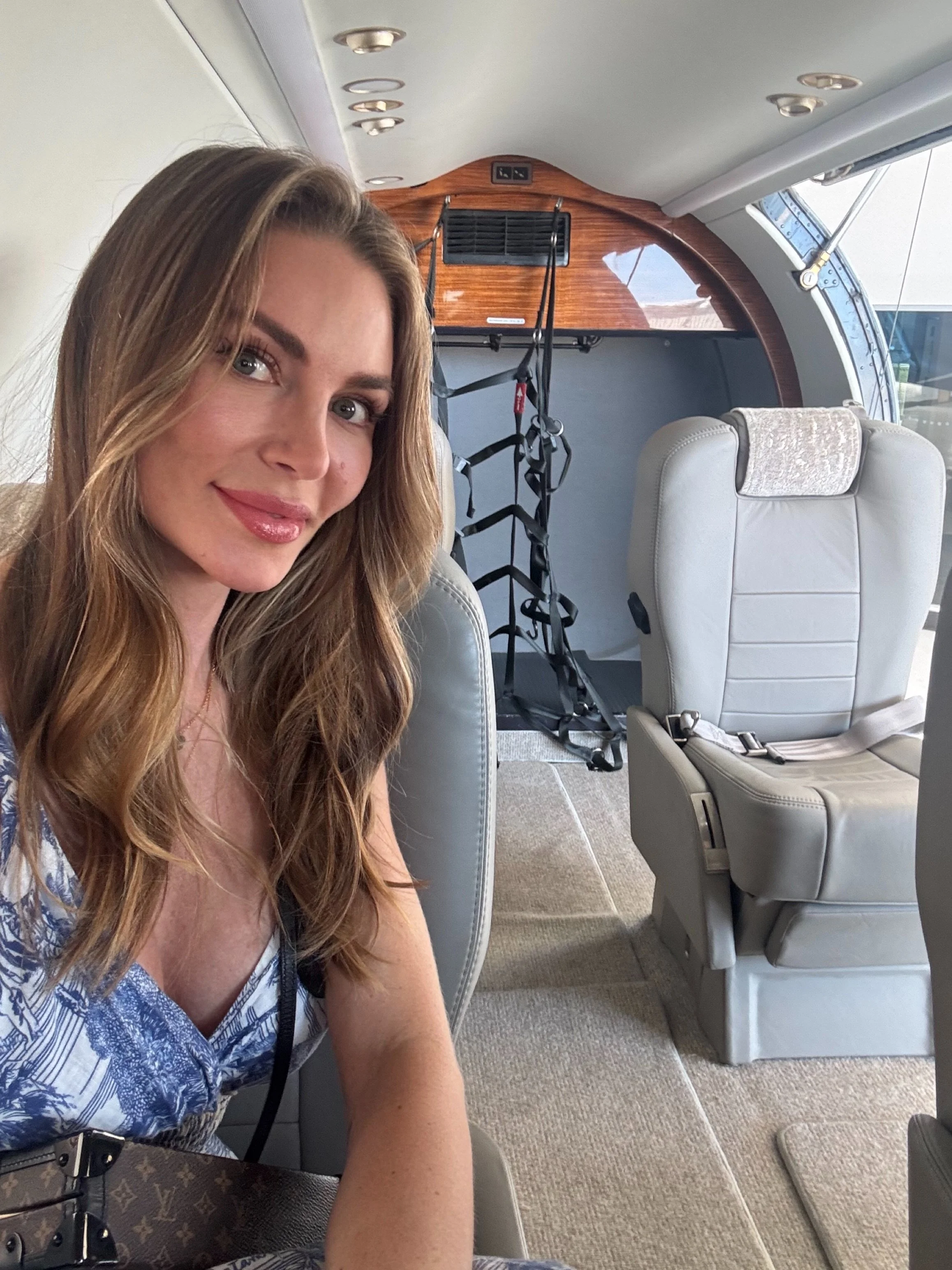A woman with long wavy hair smiling while sitting in an aircraft cabin, with empty first-class seats and a harness in the background.