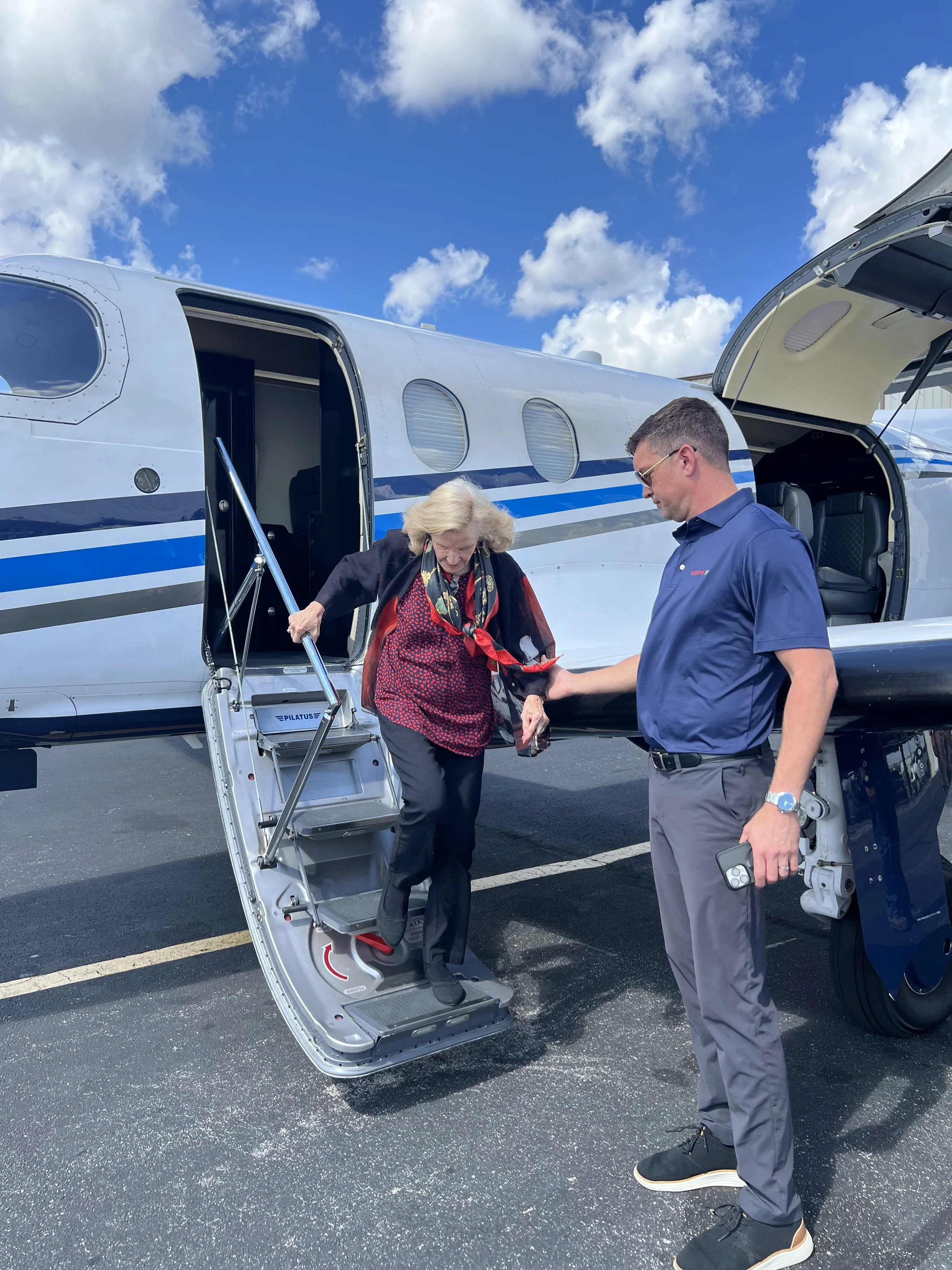 A woman is descending the stairs from a private jet, assisted by a man standing nearby on the tarmac, under a partly cloudy sky.