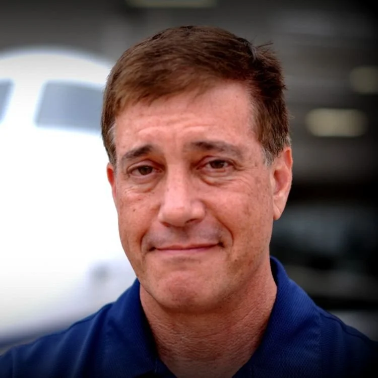 Headshot of a man with short brown hair and a slight smile, wearing a dark blue collared shirt, with a blurred airplane in the background.
