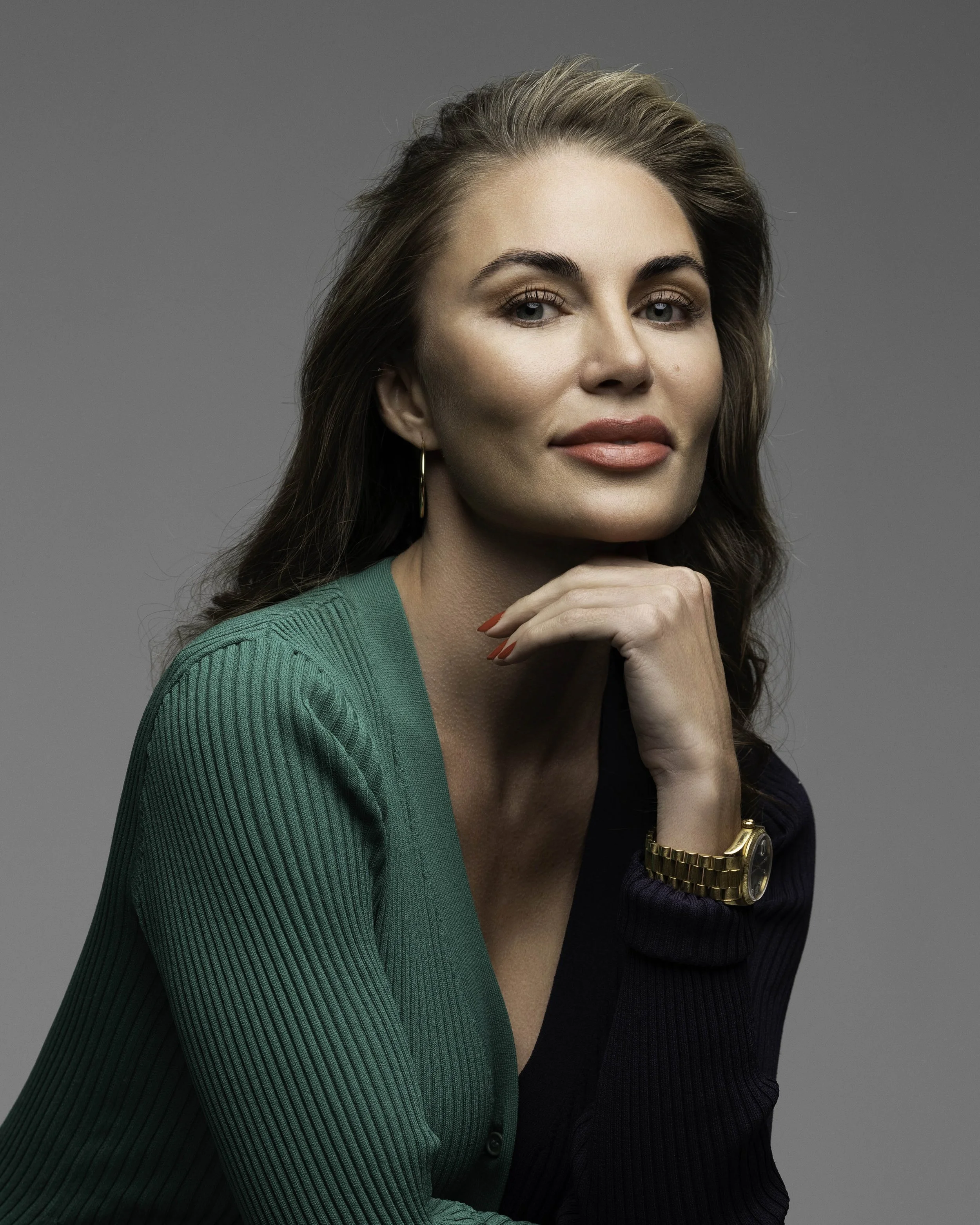 A woman with wavy brown hair, wearing a green and dark blue sweater, gold watch, and earrings, resting her chin on her hand and looking at the camera with a slight smile.
