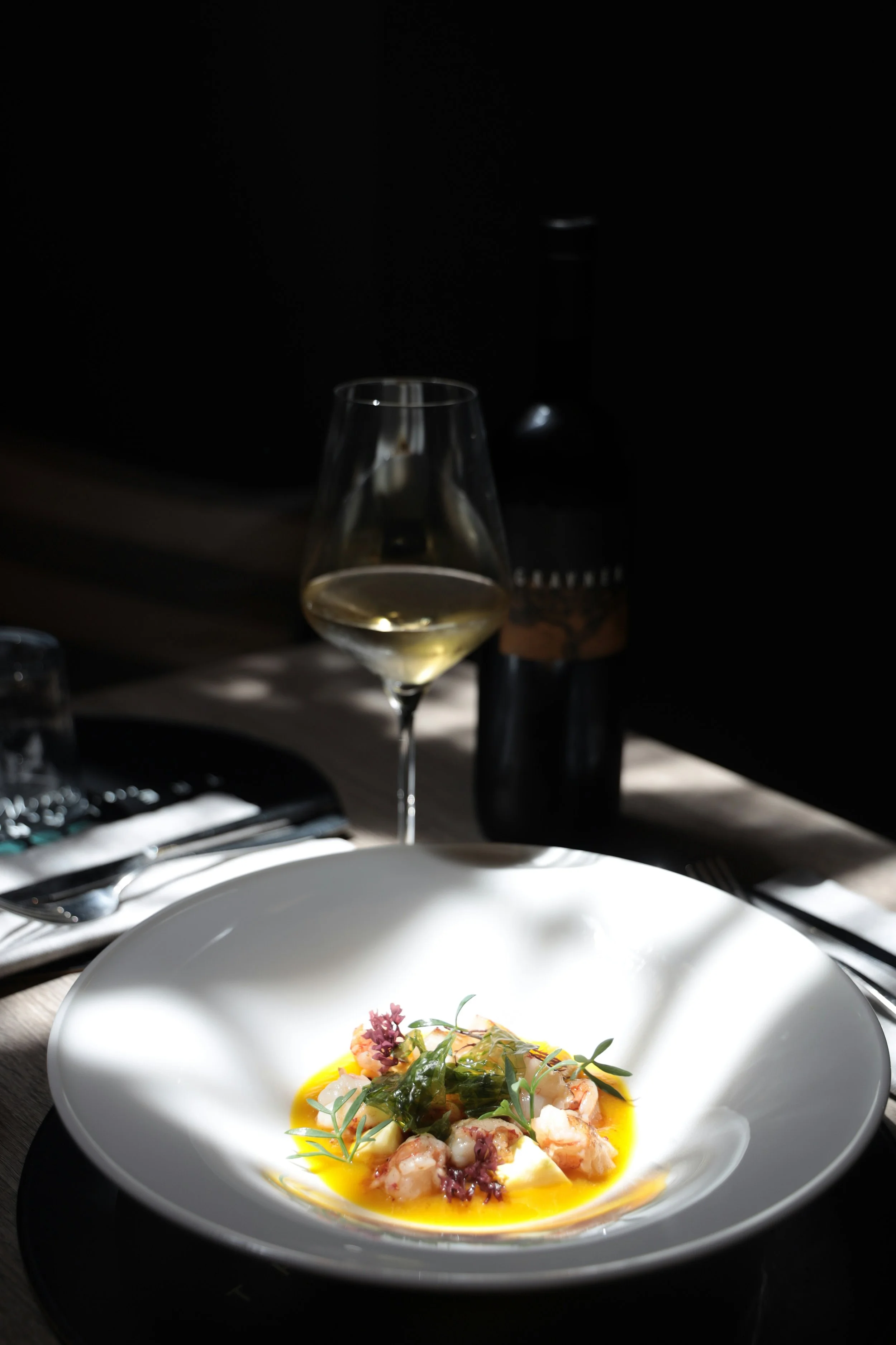 A white bowl with a gourmet seafood dish garnished with herbs and olive oil, positioned on a dark plate. Behind the dish, there is a glass of white wine with a corkscrew, a bottle of wine, and a set of cutlery on a table in a dimly lit restaurant setting.