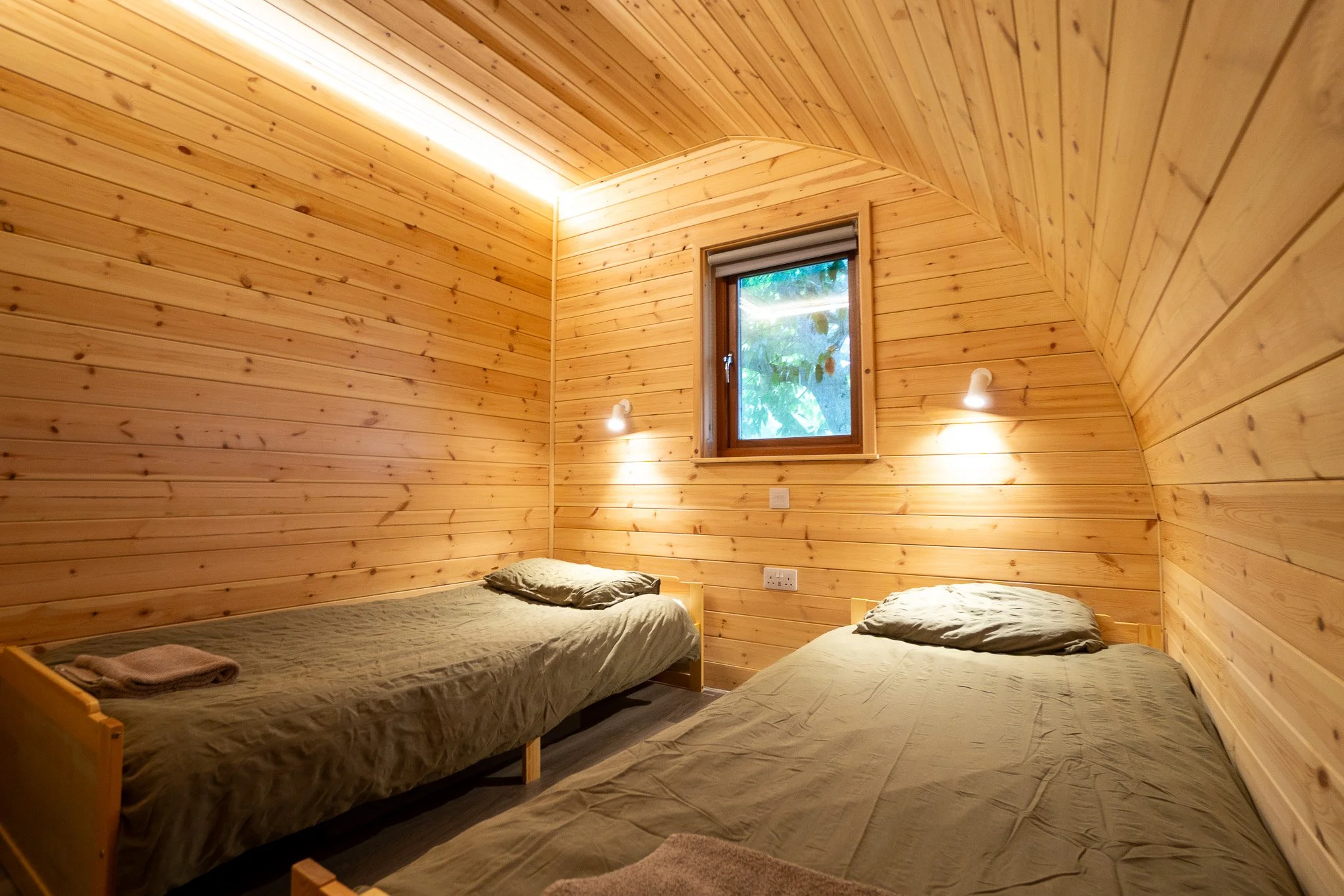 A cozy bedroom with wooden panel walls, two single beds with beige bedding, a small window with a view of greenery, and wall-mounted reading lights.