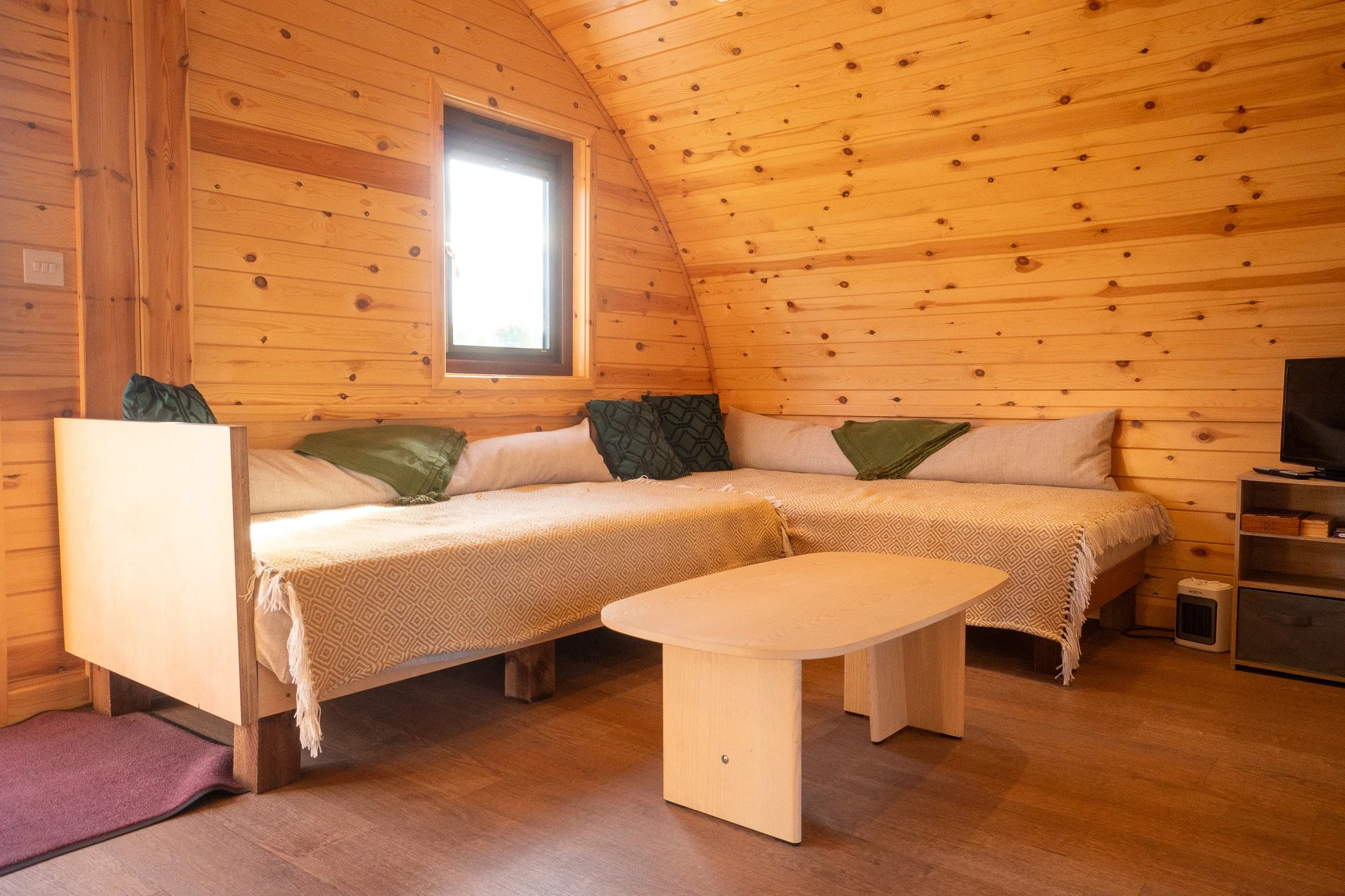 Wood-paneled interior of a cozy cabin with a large L-shaped sofa, four green pillows, a small wooden coffee table, and a television on a black stand in the corner.