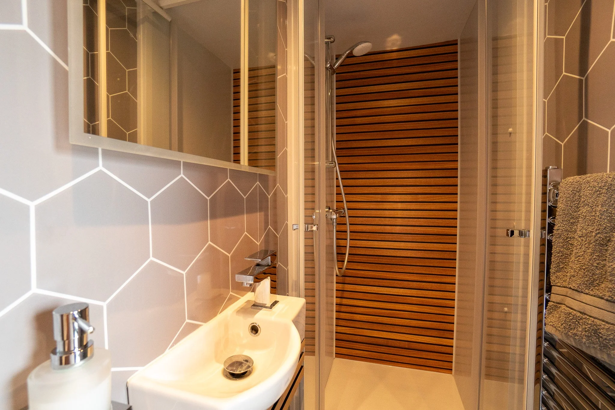 Bathroom with a small sink, soap dispenser, hexagon tiled walls, and a shower with a wooden slat wall.