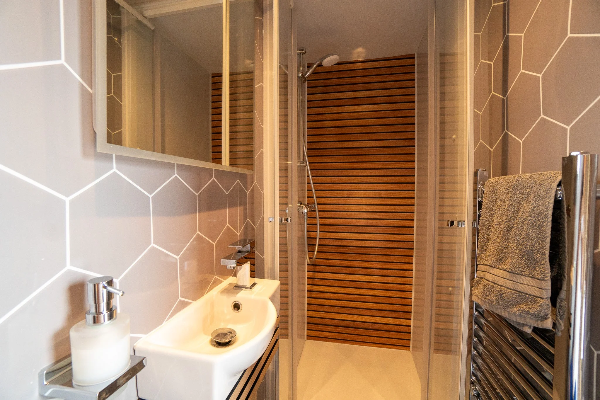 Modern bathroom with hexagonal wall tiles, a small sink, a soap dispenser, a towel hanging on a heated towel rack, and a walk-in shower with wooden panels in the background.