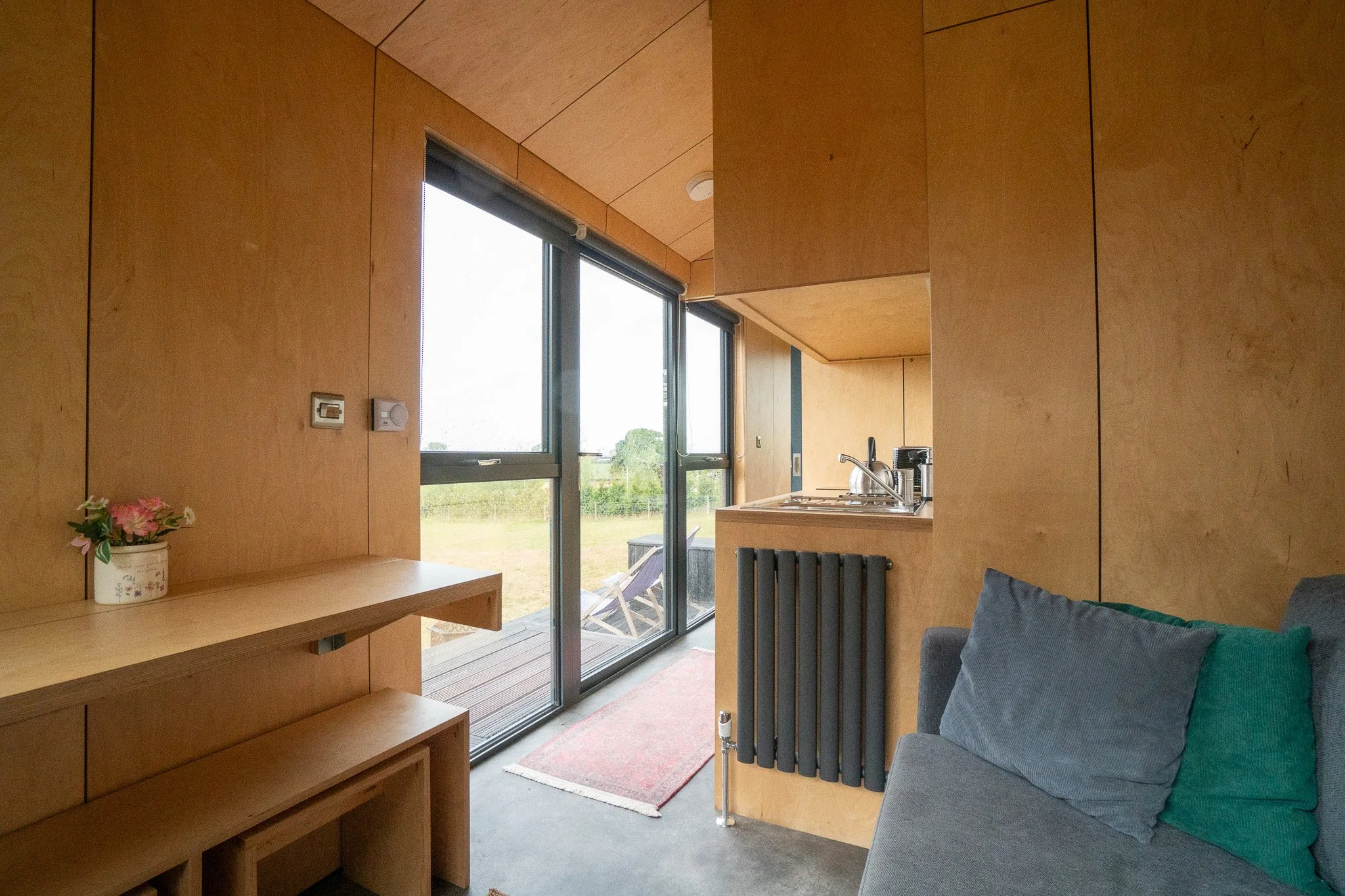 Small living space with wood-paneled walls, a gray couch with cushions, a wall-mounted black radiator, a kitchenette with a sink, and large sliding glass doors leading to an outdoor deck with a lawn chair, overlooking a grassy field.