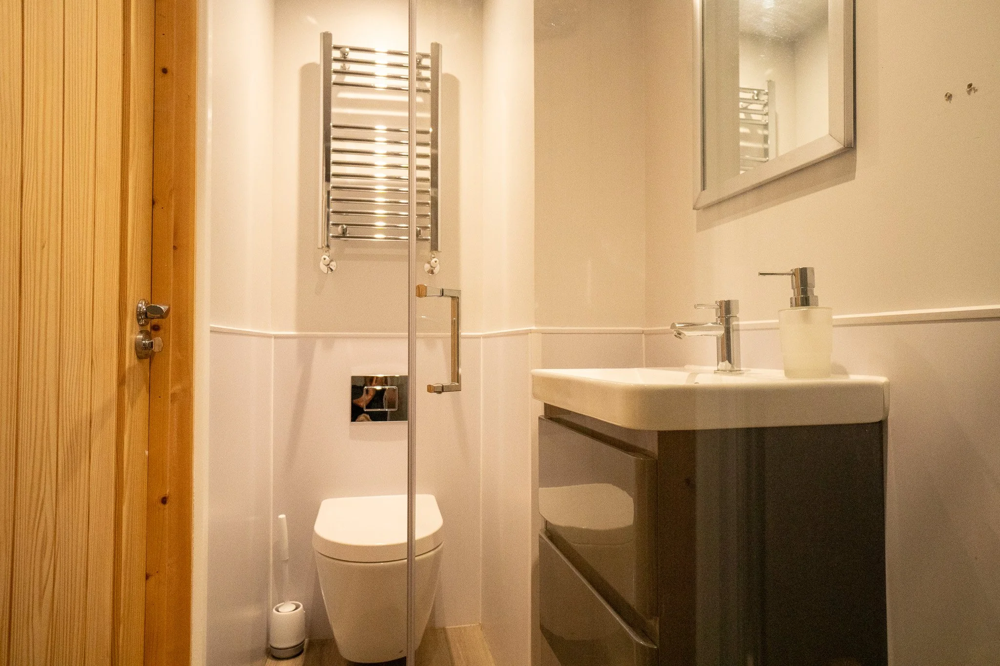 Small bathroom with white walls, a wooden door, a modern toilet, a mirror above a white sink with a faucet, and a metal towel radiator on the wall.