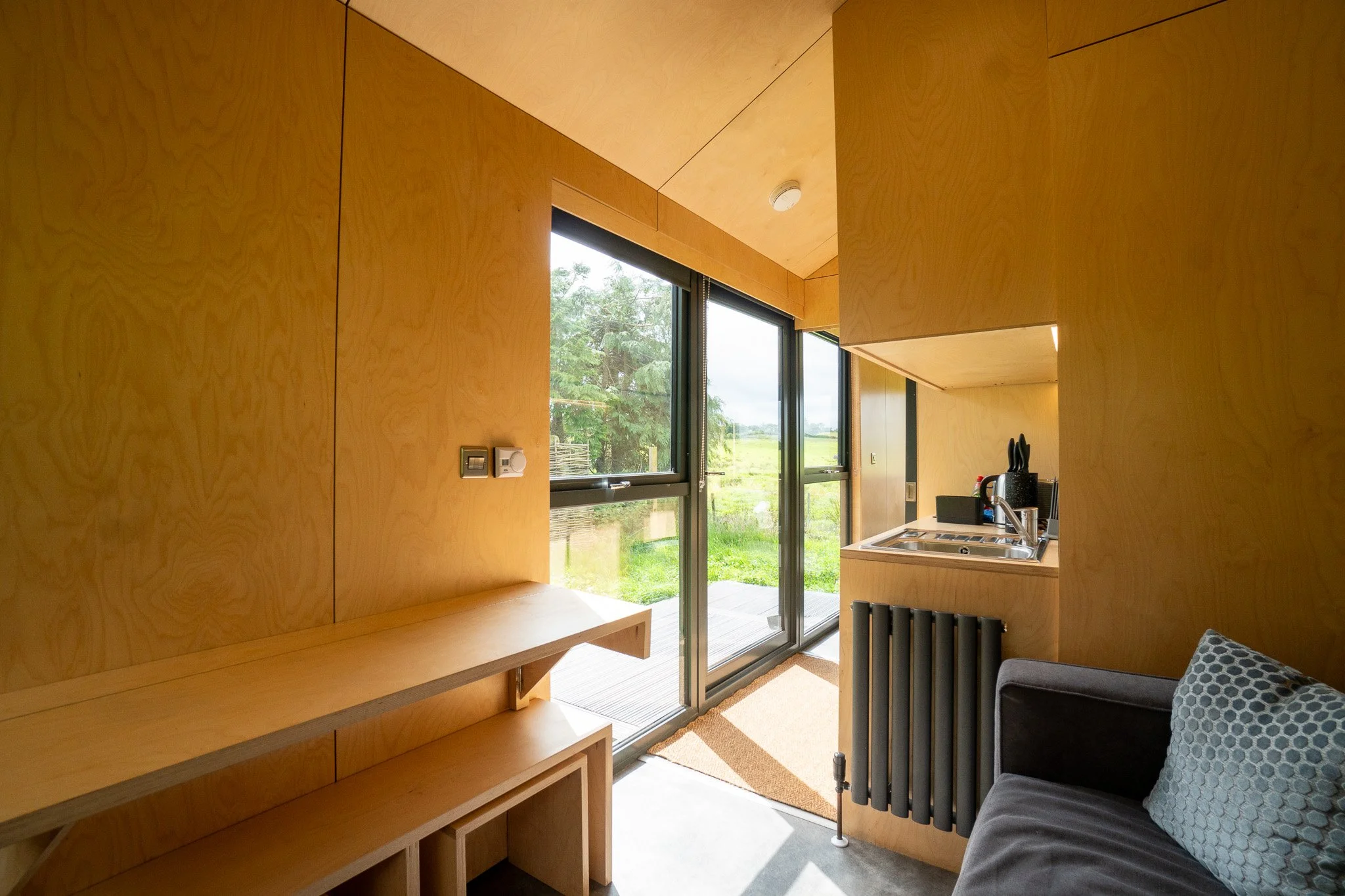 Small modern kitchen with wooden walls and large window/door overlooking green outdoor landscape, with a sofa and cushion visible in the room.