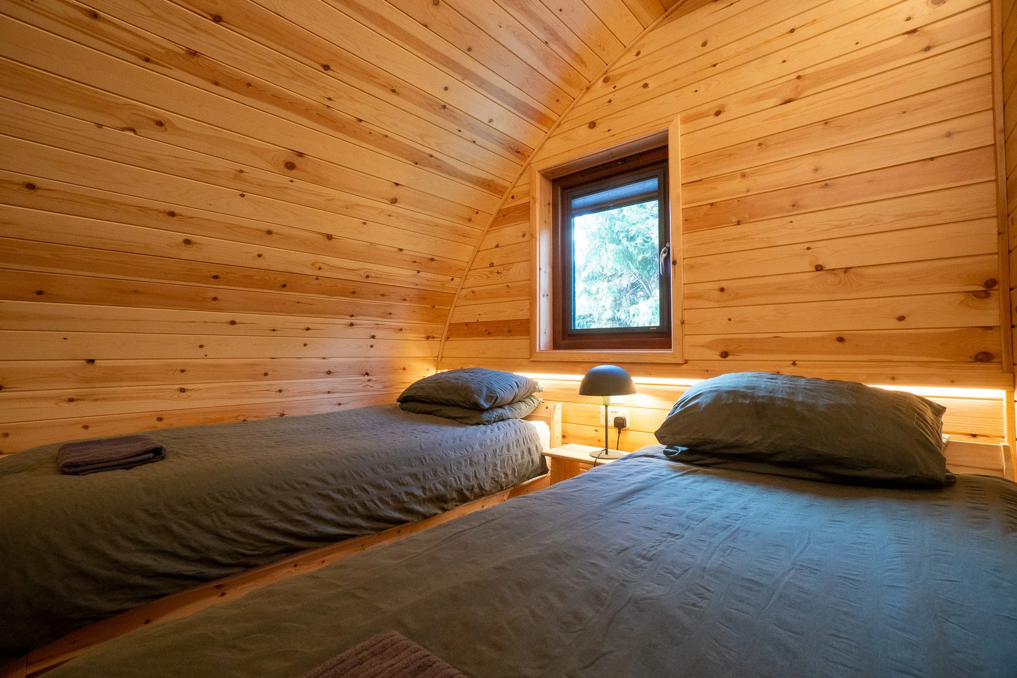 Rustic bedroom with two twin beds covered in gray bedding, wooden walls and ceiling, a window with a view of greenery, and a small bedside table with a lamp.