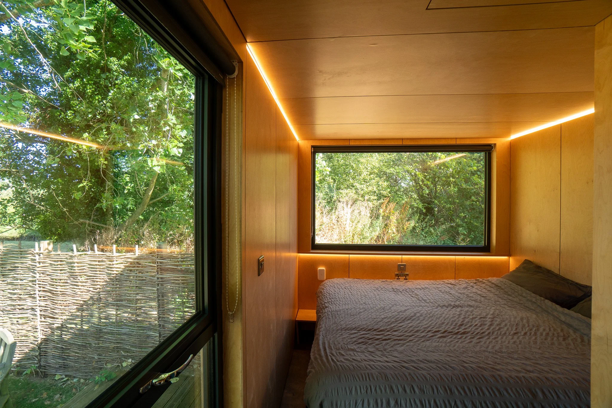 Bedroom with two large windows showing greenery outside, wooden walls, and a bed with a textured gray blanket.