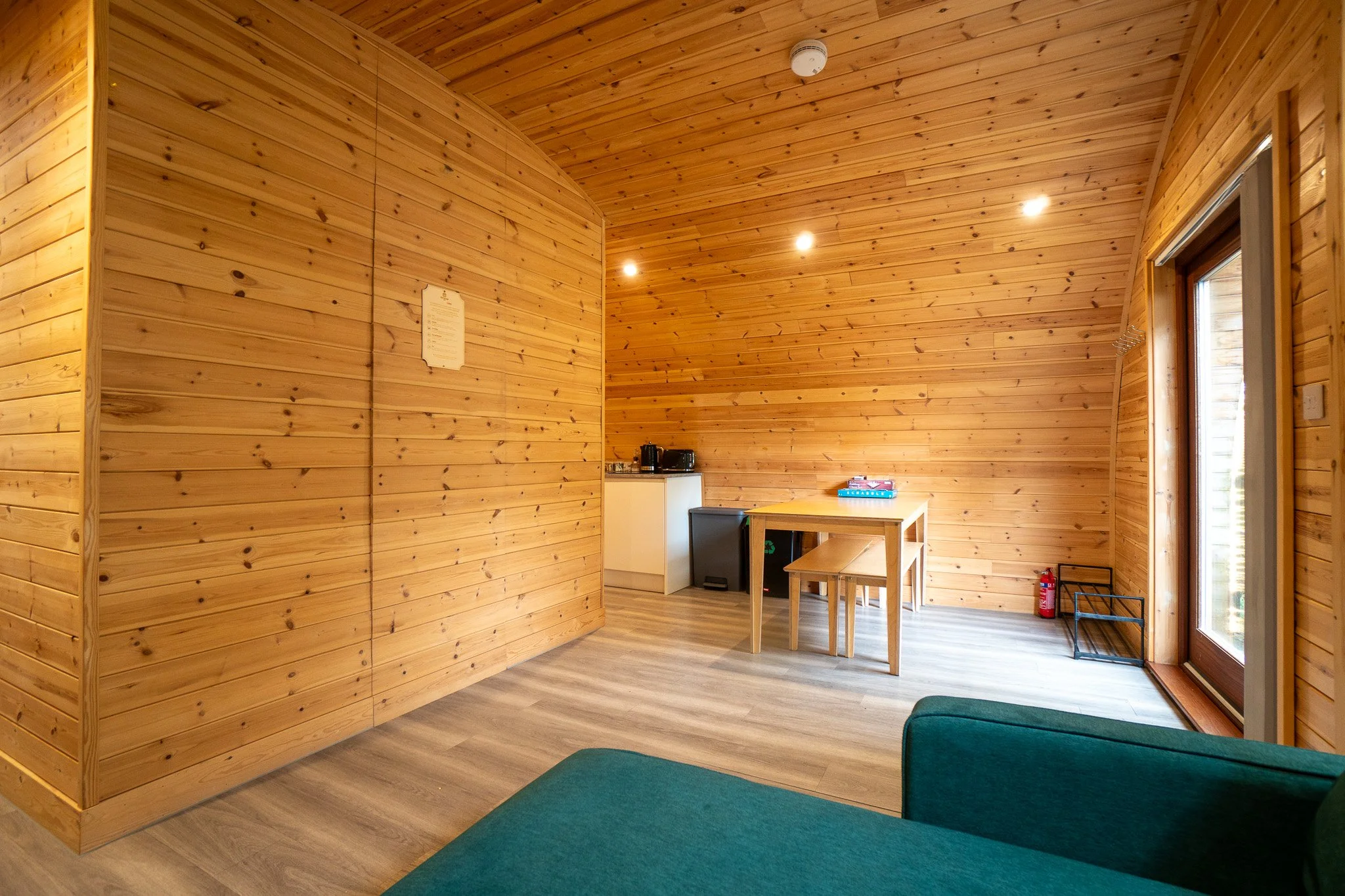 Interior of a wooden room with natural wood paneled walls and ceiling, a small dining table with two benches, a shelf with a fire extinguisher, and a sliding glass door leading outside.