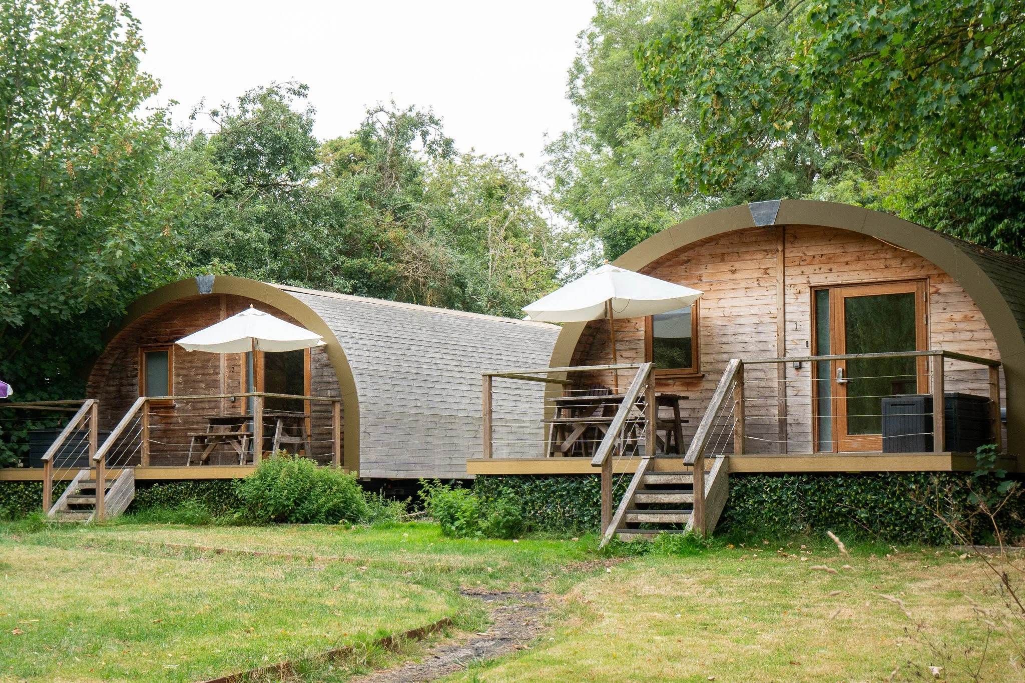 Two wooden tiny houses with curved roofs on a grassy area, each with a deck, white umbrellas, and outdoor furniture, surrounded by green trees.