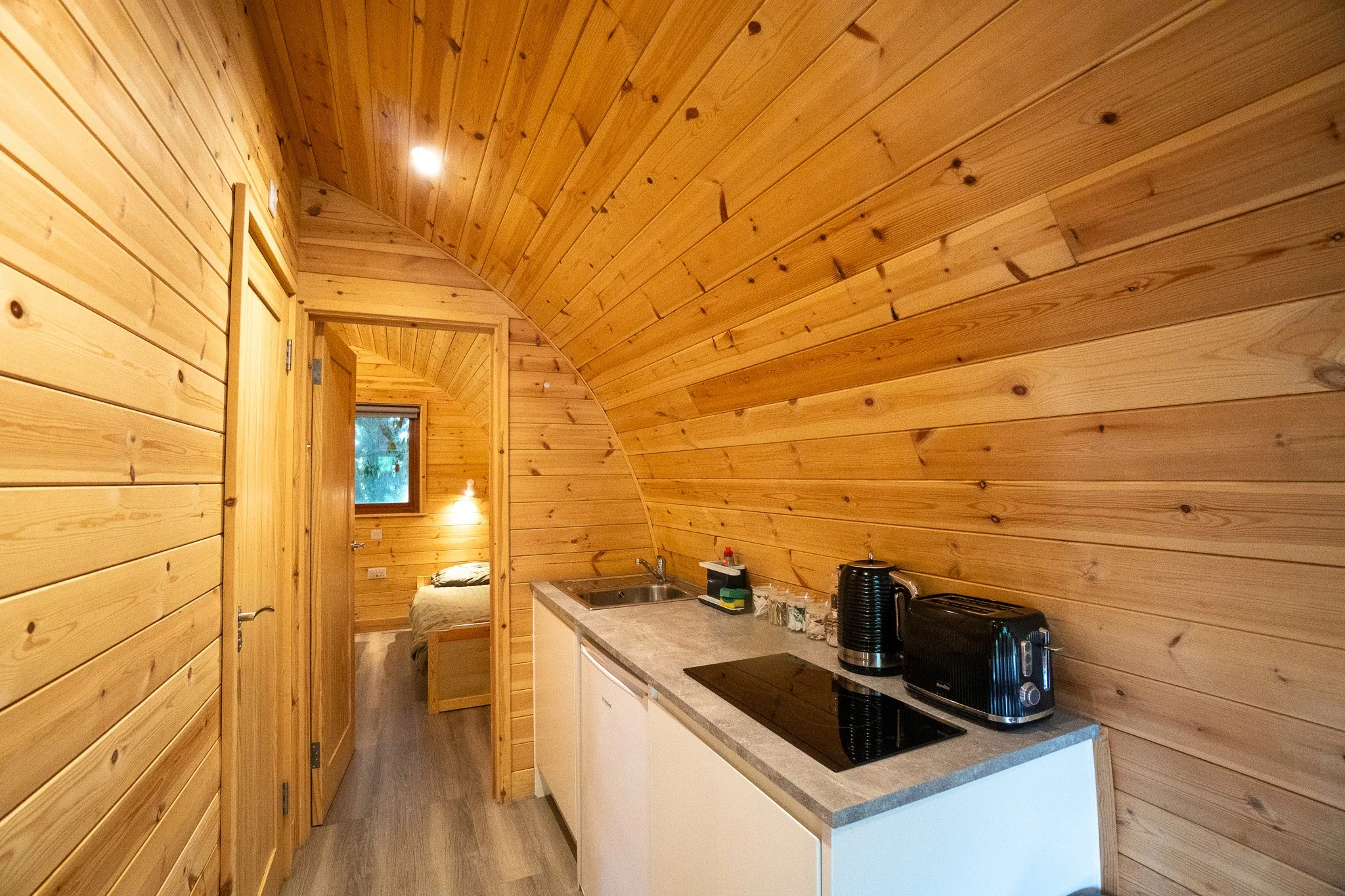 Wood-paneled kitchen with a countertop, sink, electric stovetop, kettle, toaster, and a view into a bedroom with a bed and a window.