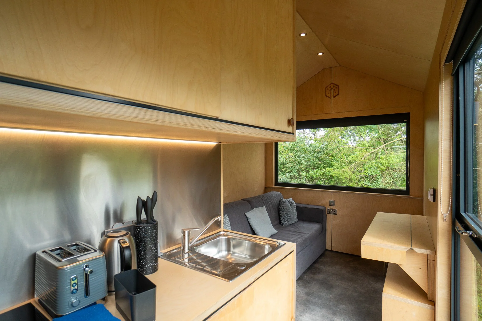 Interior of a tiny wooden kitchenette with a black sofa, a large window showing green trees, a countertop with a sink, toaster, kettle, and utensils, and a small wooden table.