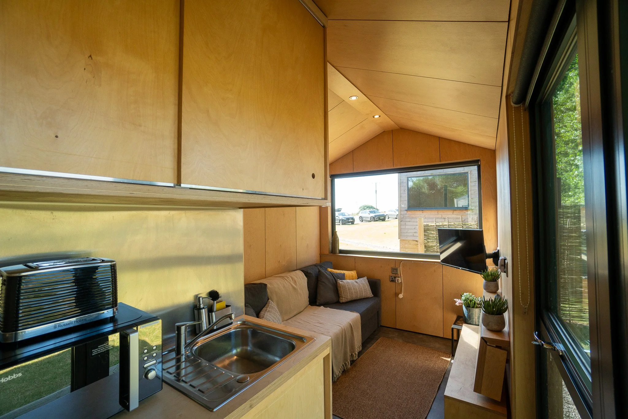 Small living space with kitchenette, couches, and a large window showing parked cars outside.