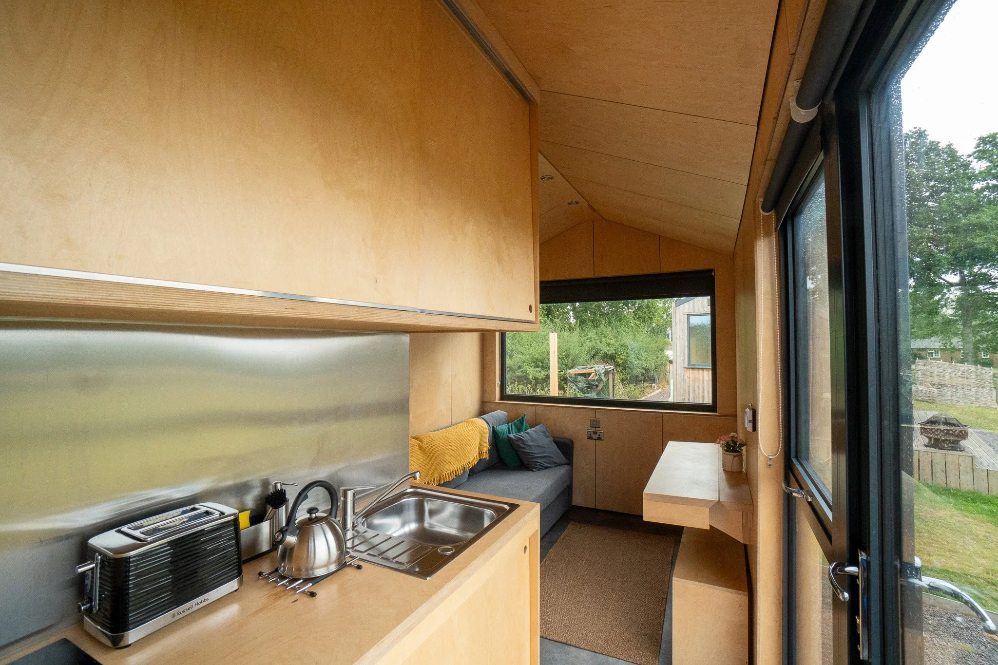 Compact wooden interior with small kitchen area, sink, toaster, and kettle; built-in bench with cushions near large window and sliding glass door leading outside.