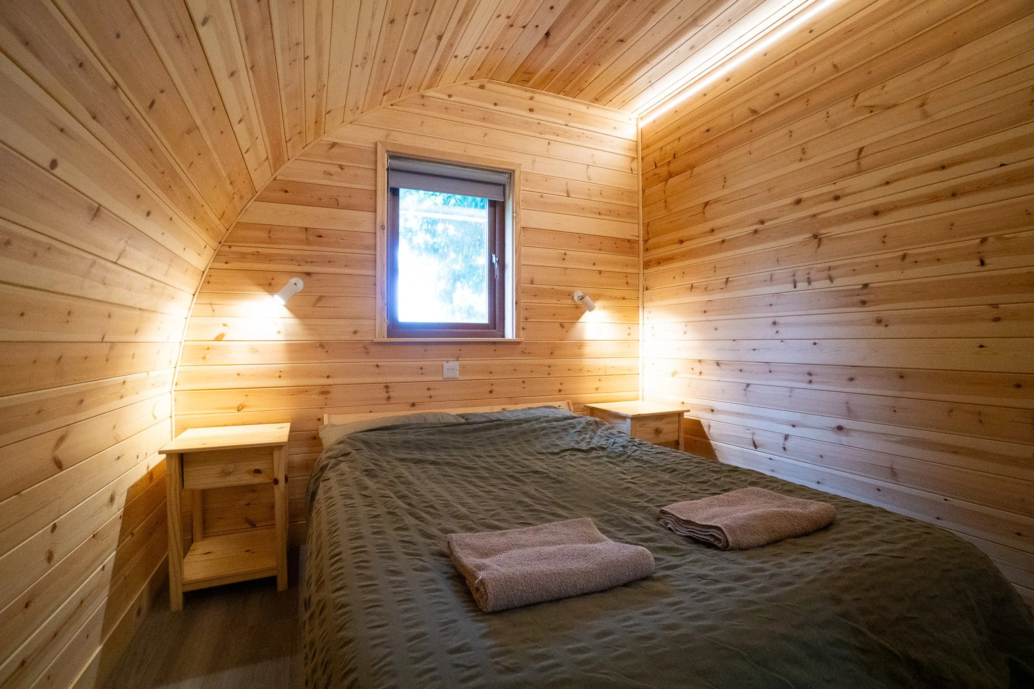 A cozy bedroom with wooden panel walls, a small window, a bed with a brown bedspread, and two towels on top. There are two small nightstands and wall-mounted reading lights.