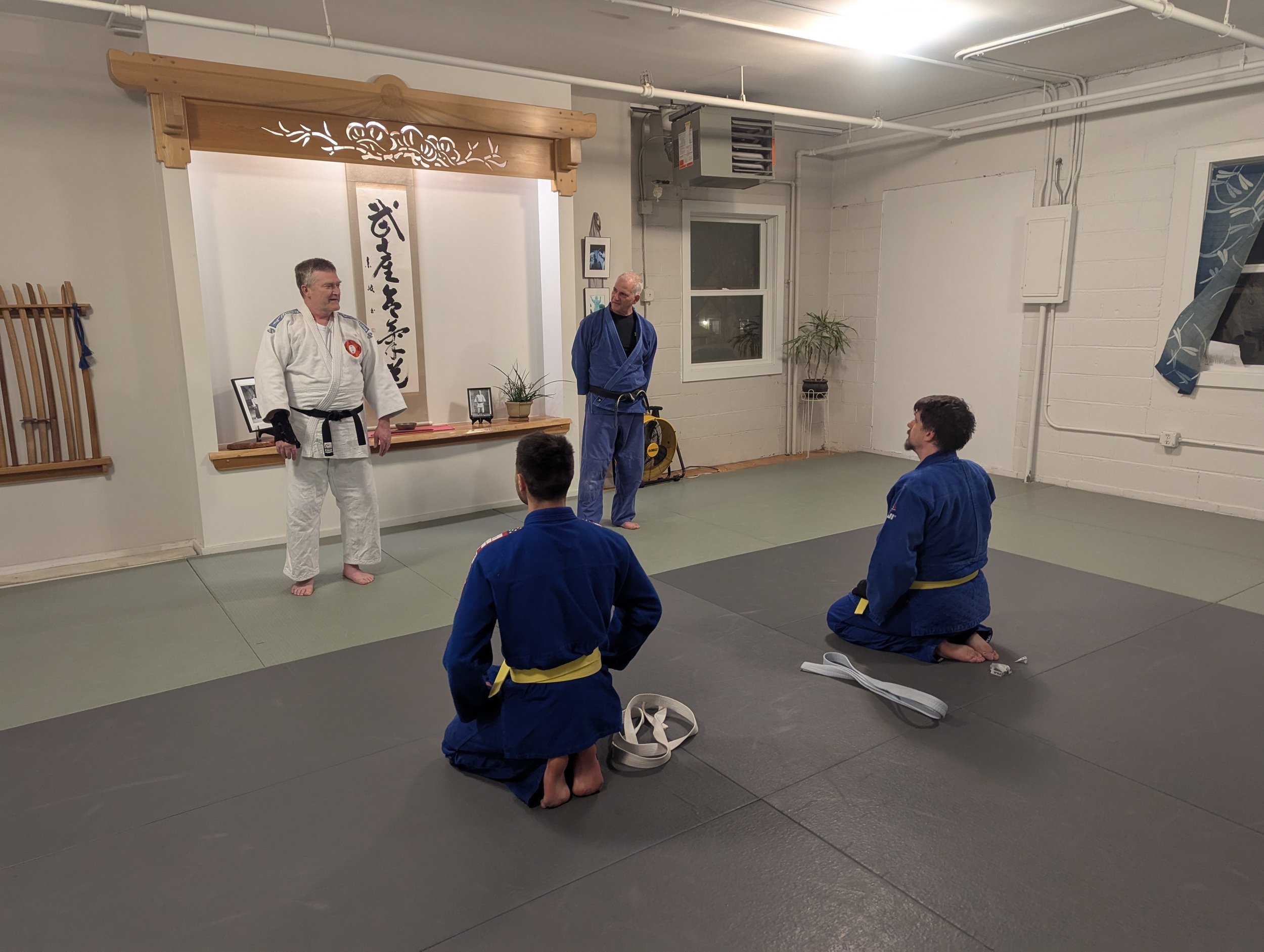 Two individuals in blue gis kneeling on the mat, facing two individuals standing in gi uniforms, one in white and one in blue, in a karate dojo with traditional decor and calligraphy