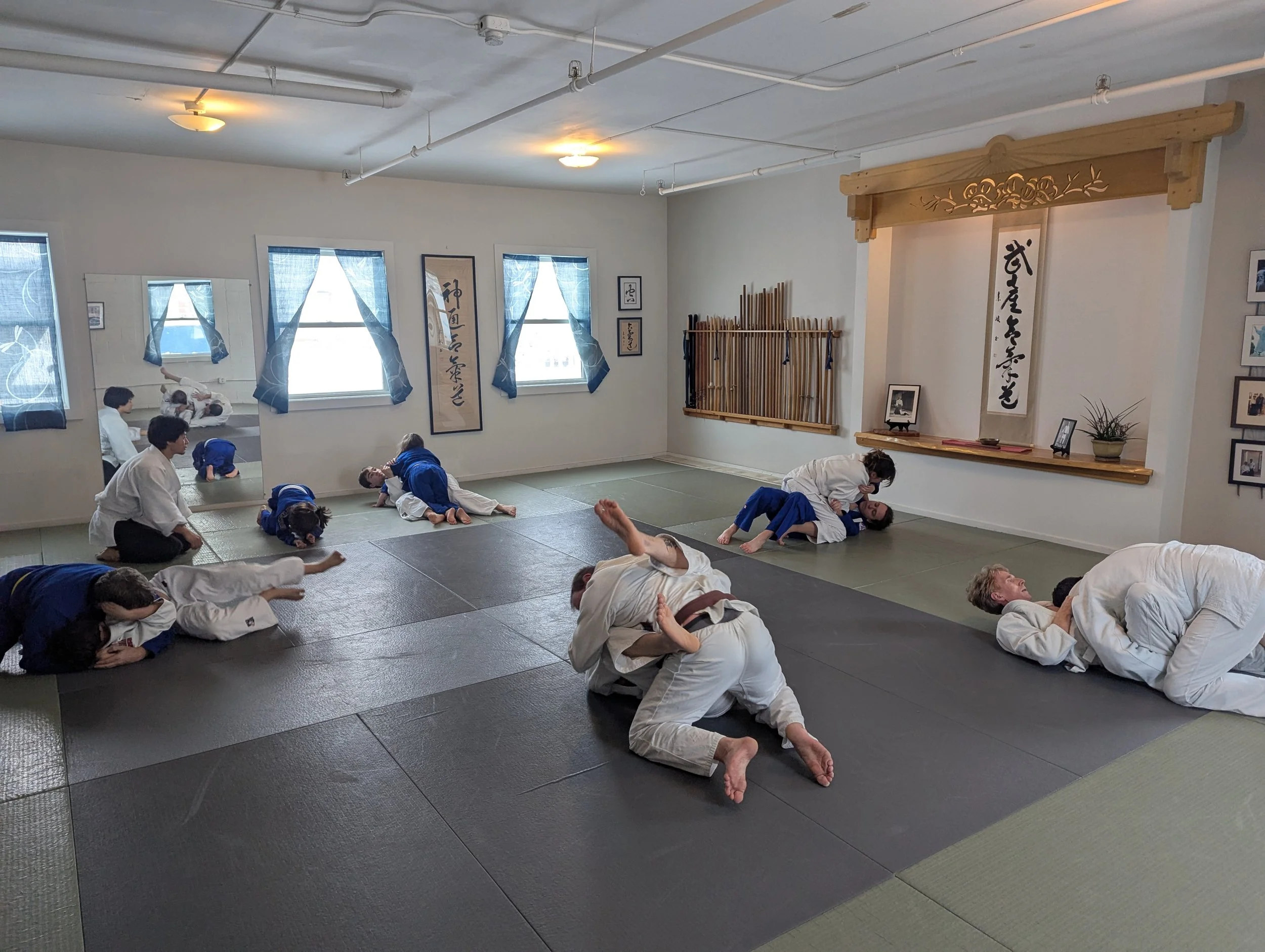 Judo students practicing techniques on mats in a dojo with instructors observing, and traditional Japanese decorations and calligraphy on the walls.