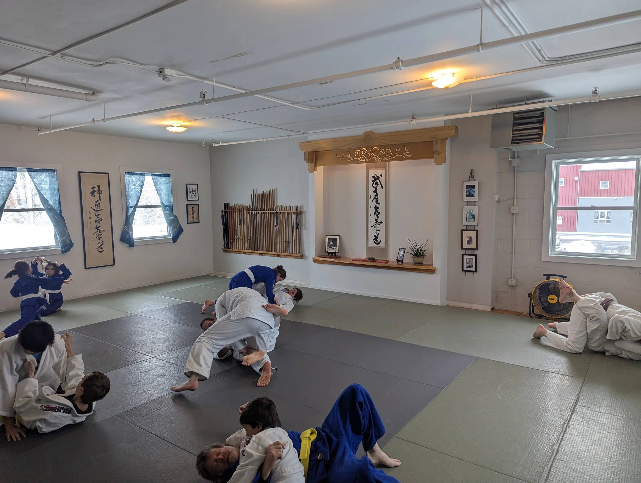 Students practicing Brazilian Jiu-Jitsu in a dojo with traditional Japanese decorations, mats on the floor, and windows allowing natural light.