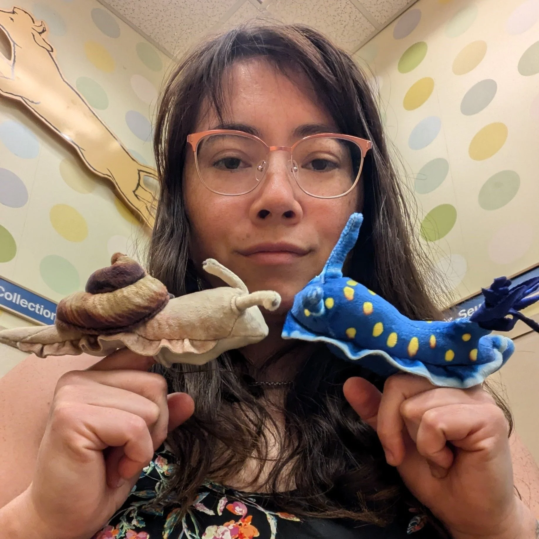 Woman with glasses holding plush snail and plush blue creature with yellow spots, in an indoor setting with polka dot wallpaper.