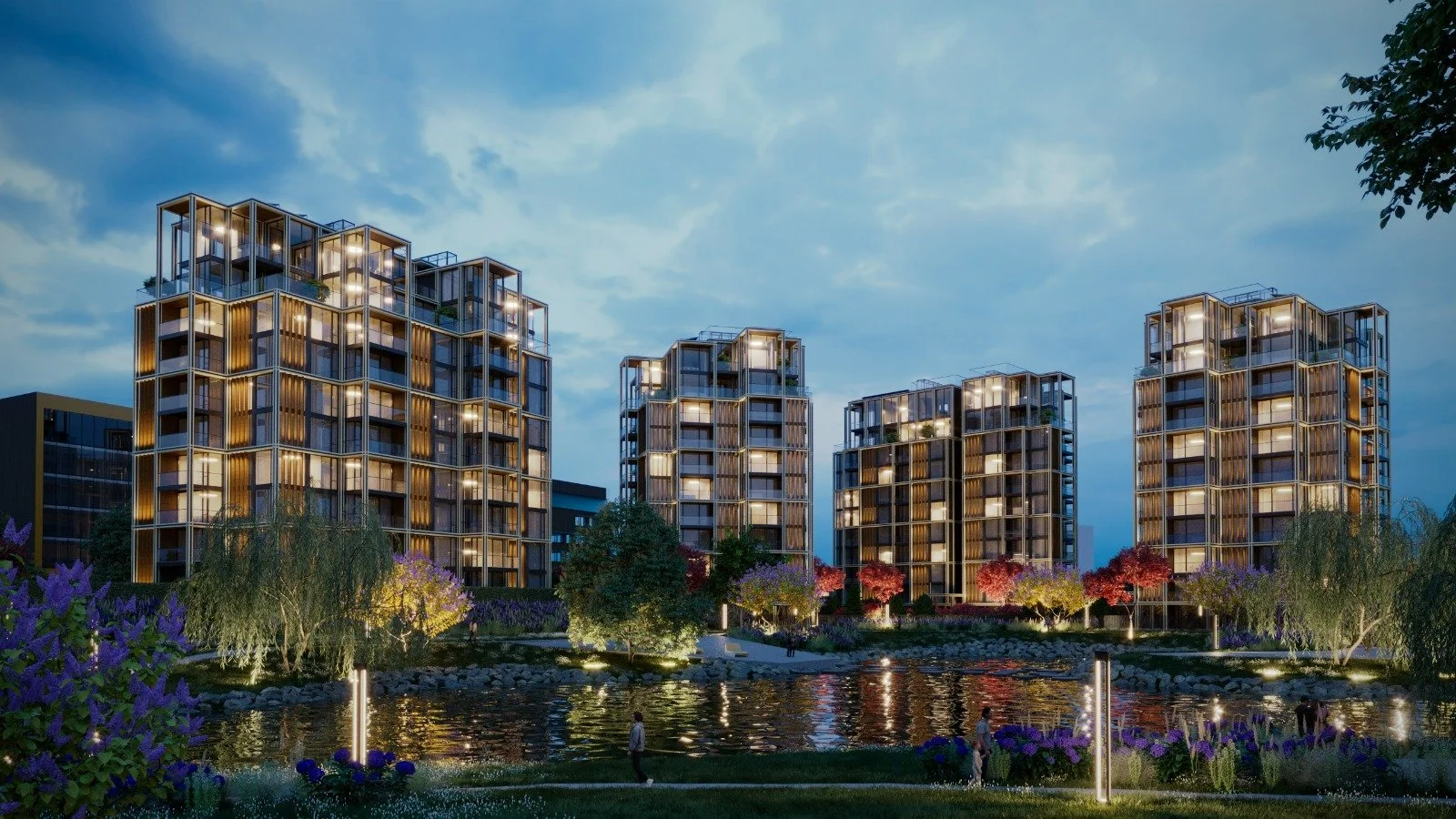 Modern apartment buildings with illuminated windows, overlooking a pond with trees and colorful flowers, under a evening sky.