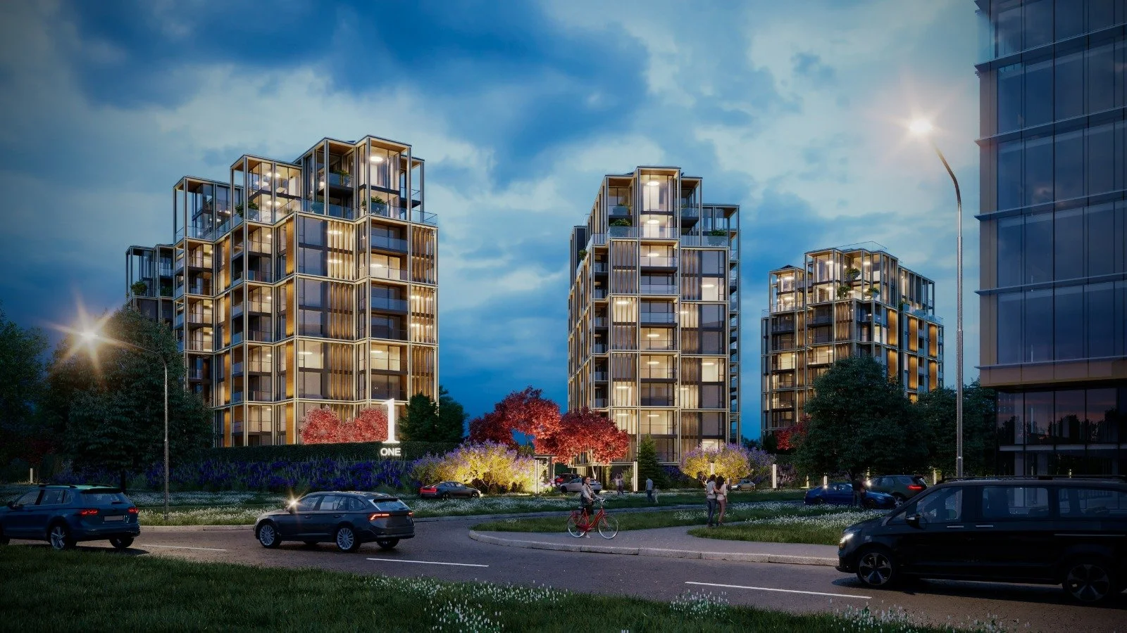 Nighttime view of a modern residential complex with several illuminated multi-story apartment buildings, trees, and cars parked along the street.