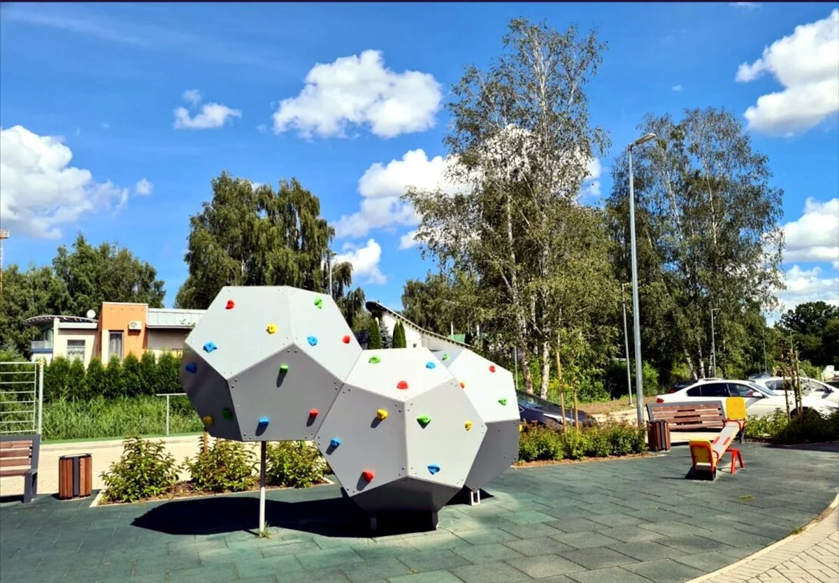 Outdoor playground with geometric climbing structures, benches, trees, parked cars, and a clear blue sky with scattered clouds.