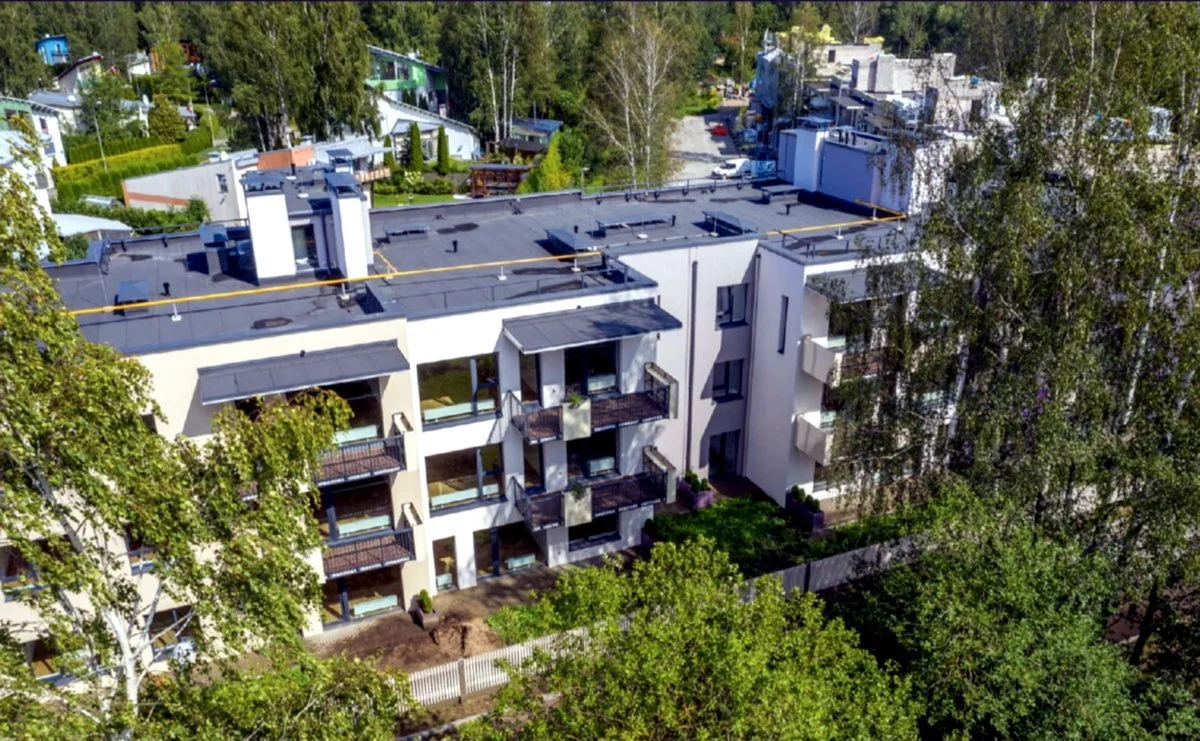 An aerial view of a modern white apartment building with balconies, surrounded by green trees and neighboring residential houses in a neighborhood.