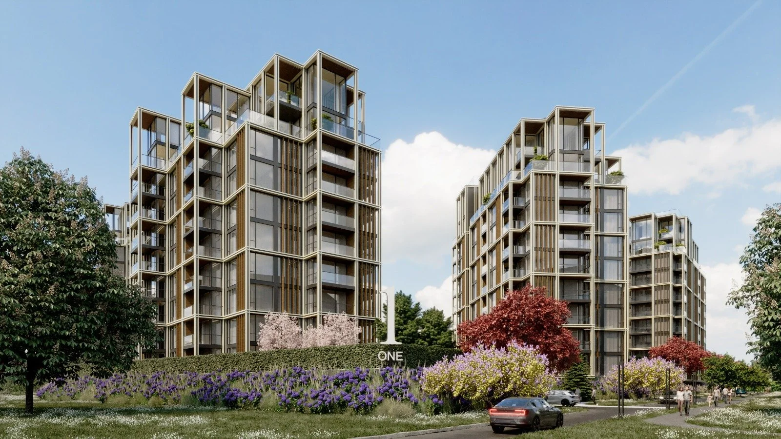 Modern multi-story residential buildings with glass balconies, surrounded by landscaped gardens with flowering trees and bushes, a pathway with pedestrians, and a dark gray car parked in the foreground under a partly cloudy sky.