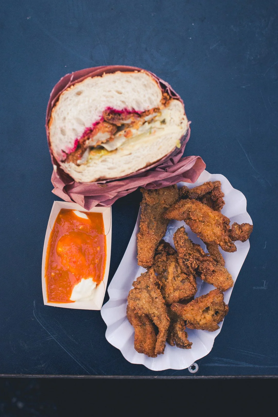 Ein Sandwich mit der Öffnung nach oben und einer Füllung im oberen Bild. Darunter liegt eine Schale mit Frittierten Stücken , daneben eine weiße Soße, die mit einer orangen Soße überzogen ist.