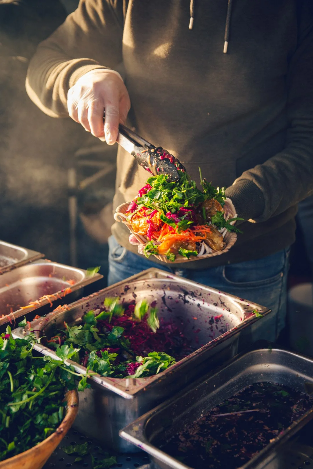 Eine Person trapiert Kräuter mit einer Zange auf einer Schale voll mit Essen. Dabei fallen einige Kräuterblätter herunter. Diese fallen direkt in die Schale zurück oder in die Schale, die davor platziert ist. Hier befindet sich ein rotes Kraut.