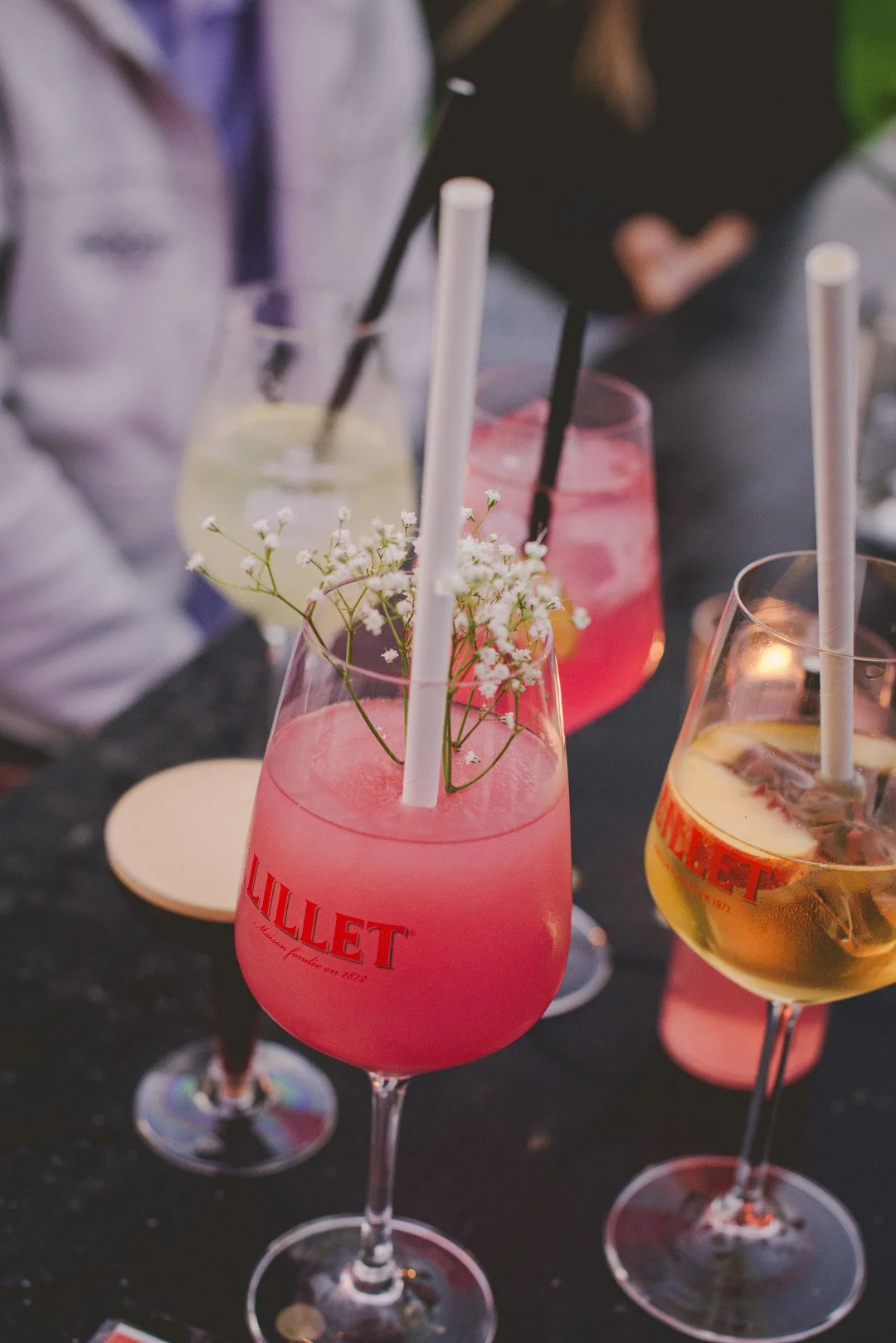 Kühle Sommerdrinks in Weingläsern stehen auf einem Tisch mit Personen im Hintergrund - rosa, orange und gelb mit frischen Früchten,Minze  und Strohhalmen.