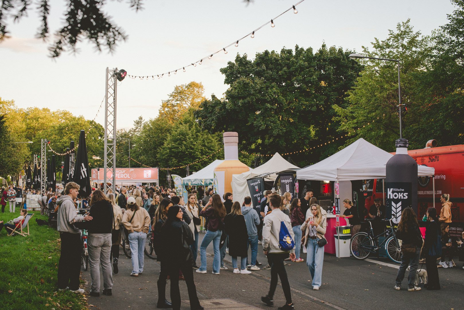 Belebtes Straßenfest mit Getränke- und Essensständen. Abendstimmung mit Lichterketten auf einer Straße am Park.