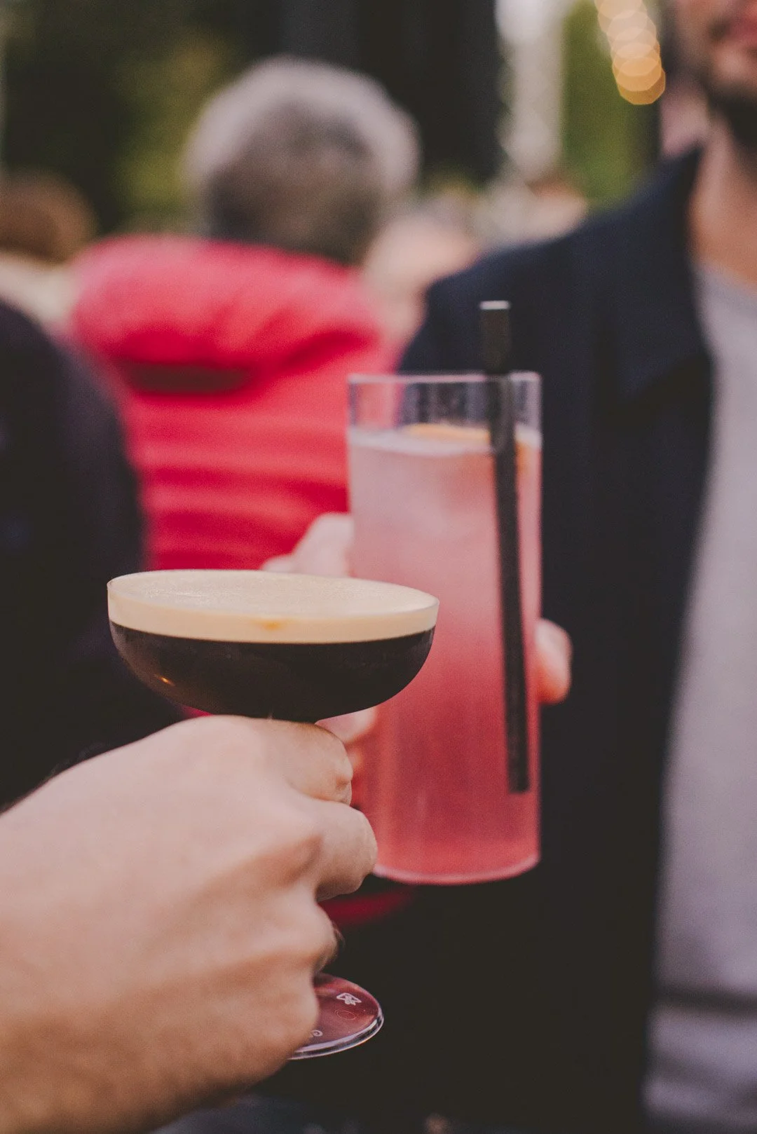 Zwei Personen stoßen an – mit einem Espresso Martini und einem rosa Spritz im Kölschglas mit Strohhalm.