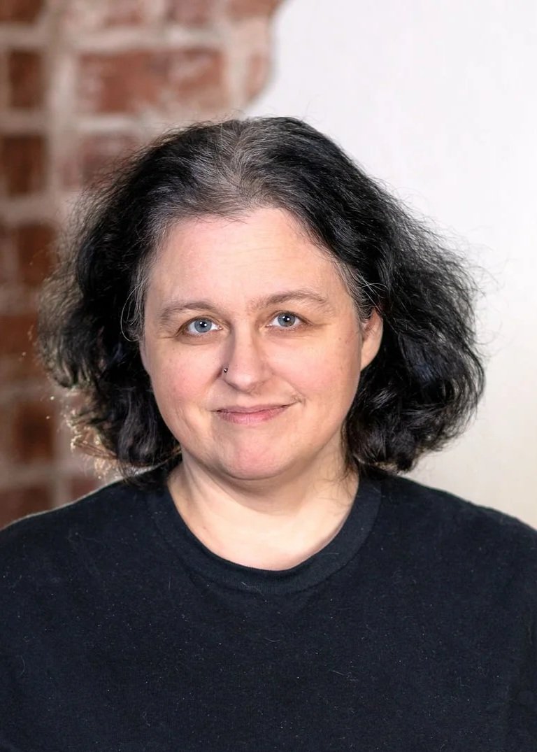 A woman with with dark hair and blue eyes, wearing a black top, against a gray wall background.